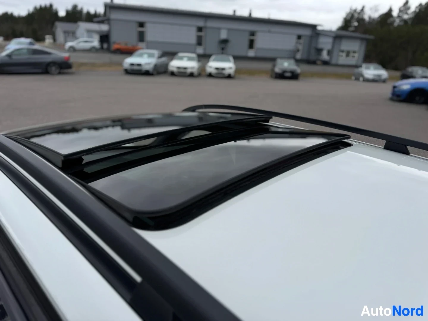 Yksityiskohta: Valkoisen auton katto, jossa on avoin panoraamakattoluukku. Taustalla näkyy matala rakennus ja muita autoja.