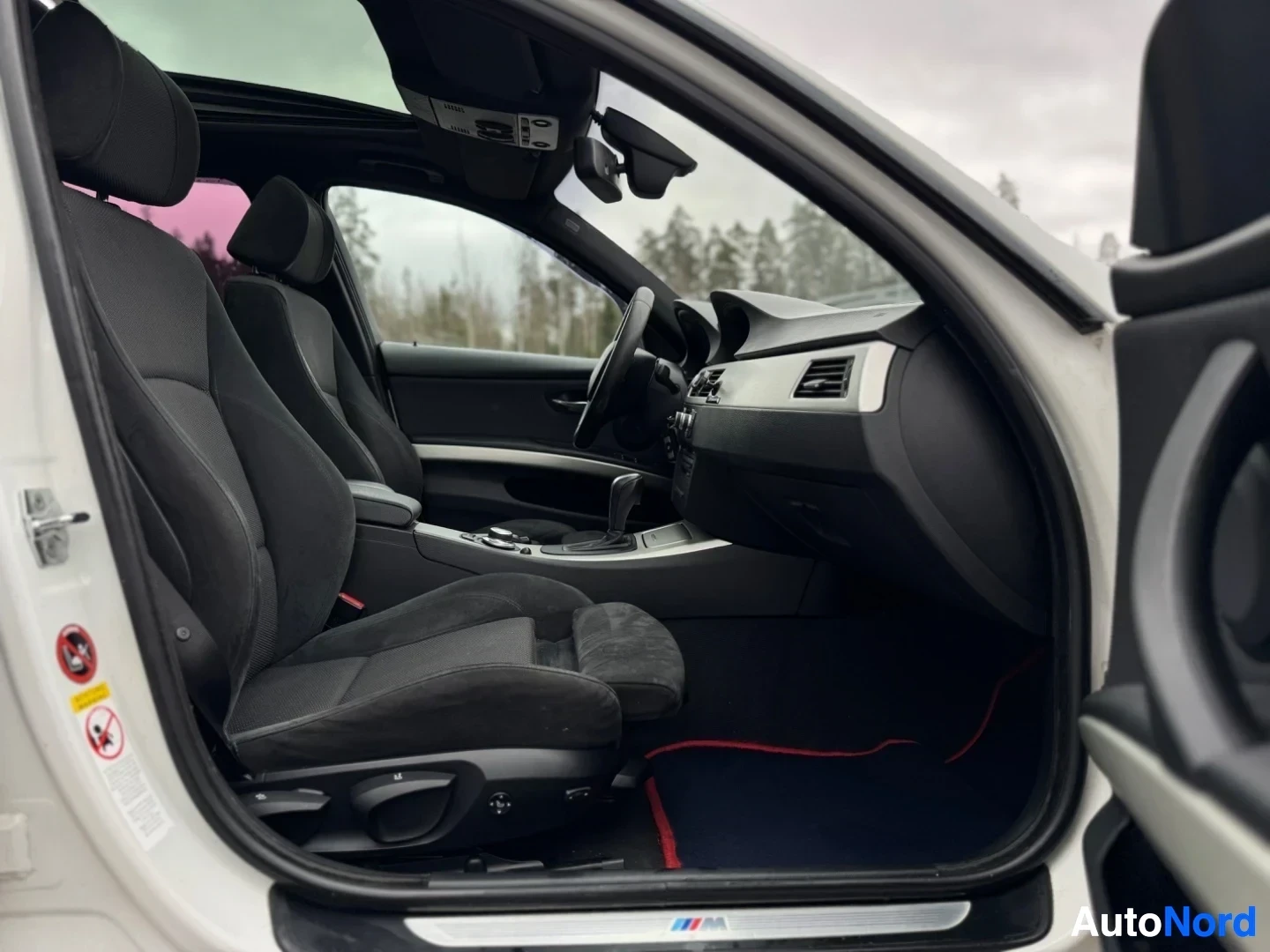 Ohjaamo: Kuva auton etuistuimista ja ohjaamosta kuljettajan puolelta, kuljettajan ovi auki. Tummat kangas/alcantara-istuimet, ohjauspyörä ja mittaristo näkyvissä. Kattoi
