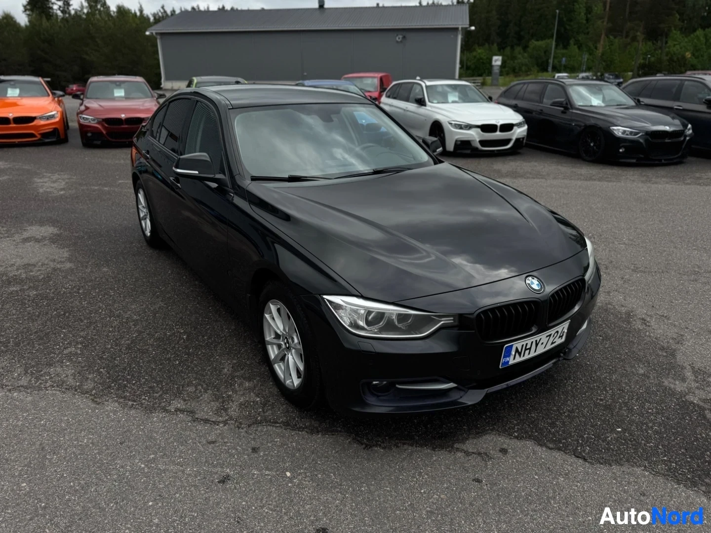 Etuviisto, nokka oikealle: Musta BMW sedan edestä kuvattuna, rekisterikilpi "" näkyvissä. Taustalla on pysäköintialue täynnä autoja ja rakennuksia