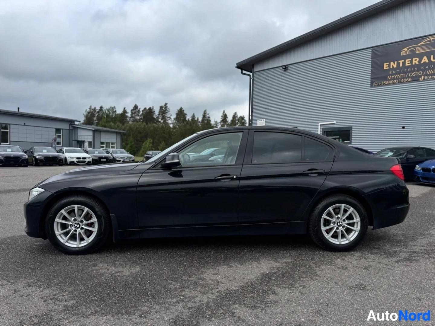 Sivulta: Musta BMW sedan sivusta kuvattuna, pysäköitynä rakennuksen eteen. Auto on taustalla olevan mainostaulun vasemmalla puolella.