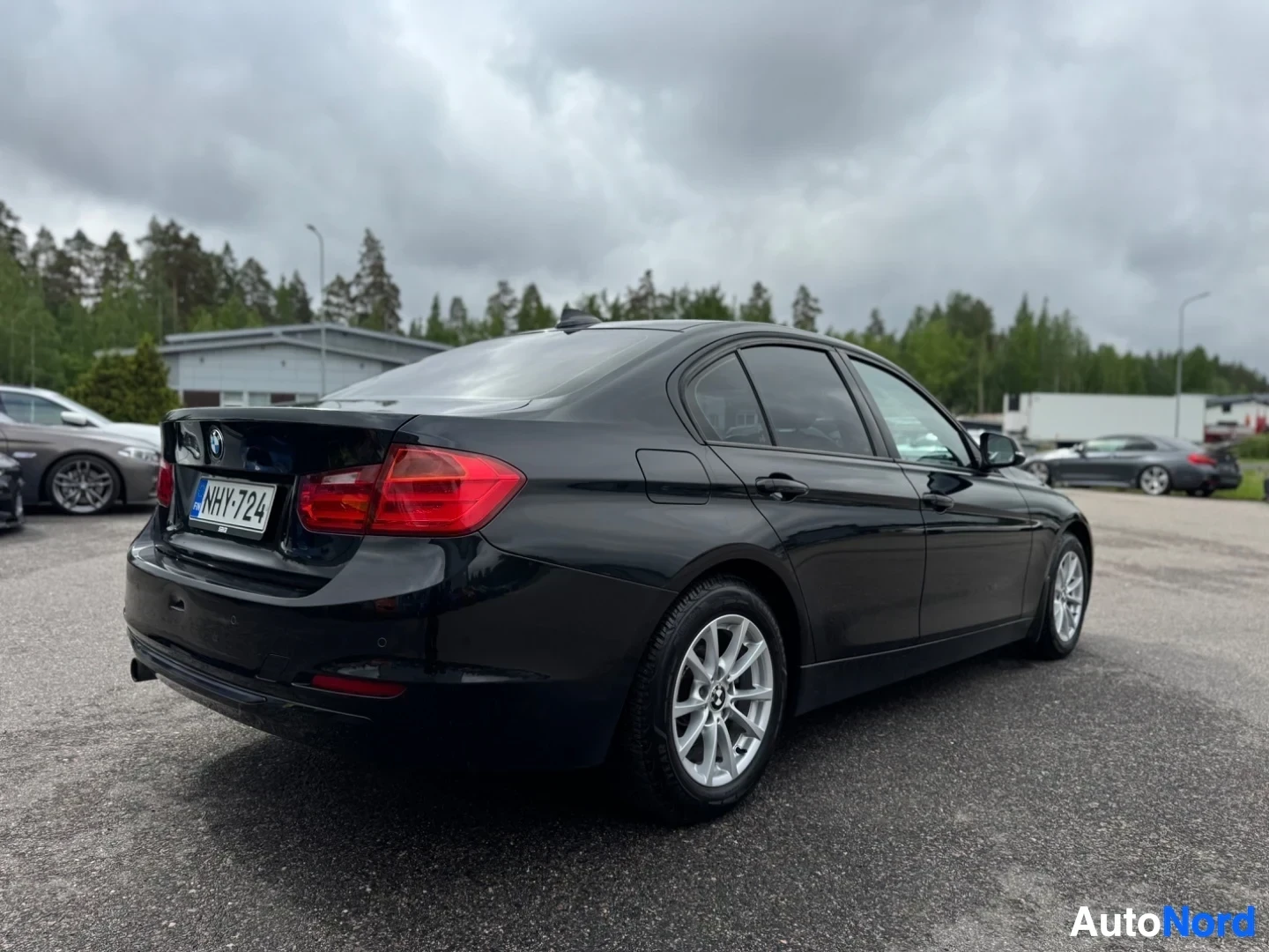 Takaviisto: Musta BMW sedan, jossa on rekisterikilpi "", pysäköitynä ulkona taustalla puita ja rakennuksia