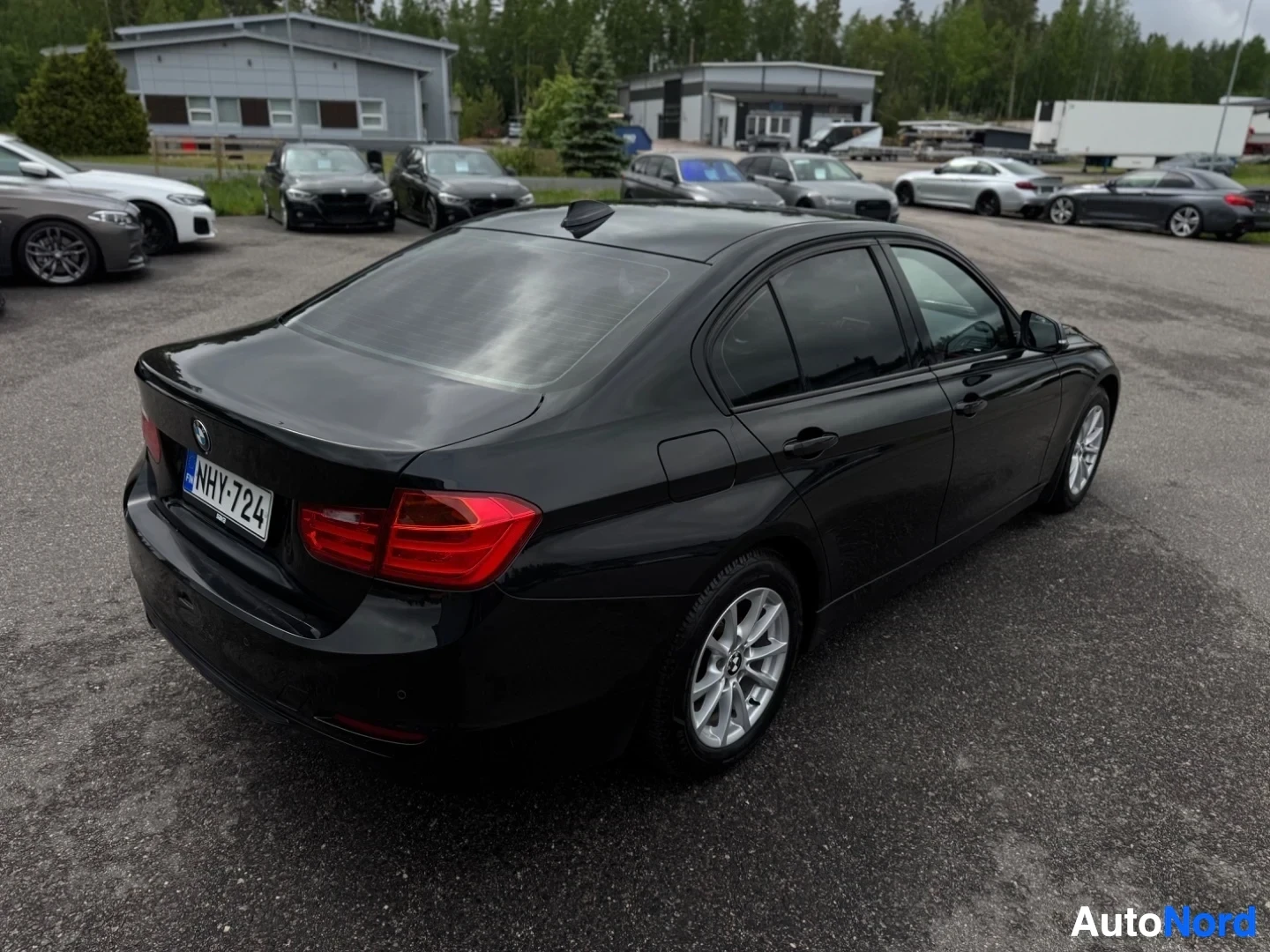 Takaviisto: Musta BMW sedan takaviistosta kuvattuna, rekisterikilpi "" näkyvissä. Taustalla on rakennuksia ja muita pysäköityjä autoja