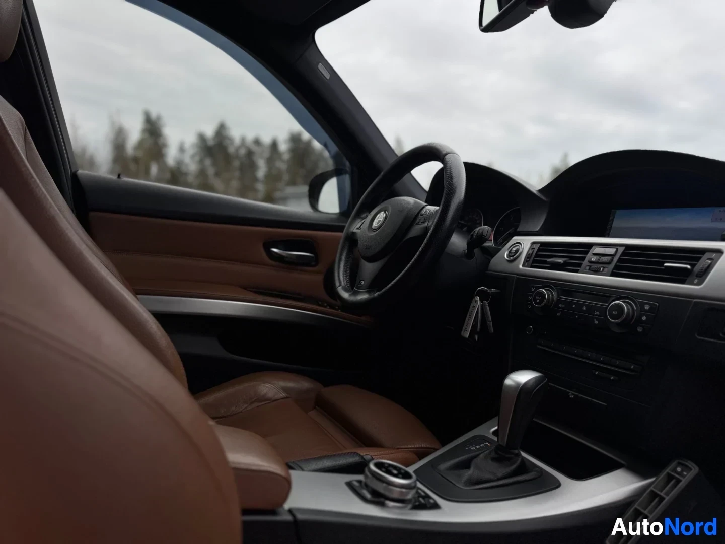 Ohjaamo: Kuvakulma kuljettajan oven puolelta auton ruskeaan nahkaverhoiltuun sisätilaan. Musta ohjauspyörä, jossa BMW-logo, näkyy, ja keskikonsolissa on näyttö ja painik