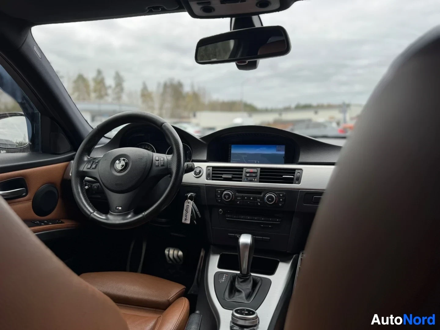Ohjaamo: Kuvakulma etumatkustajan ovesta auton ruskeaan nahkaverhoiltuun sisätilaan. Musta ohjauspyörä, jossa BMW-logo ja monitoimipainikkeet, näkyy. Keskikonsolissa on