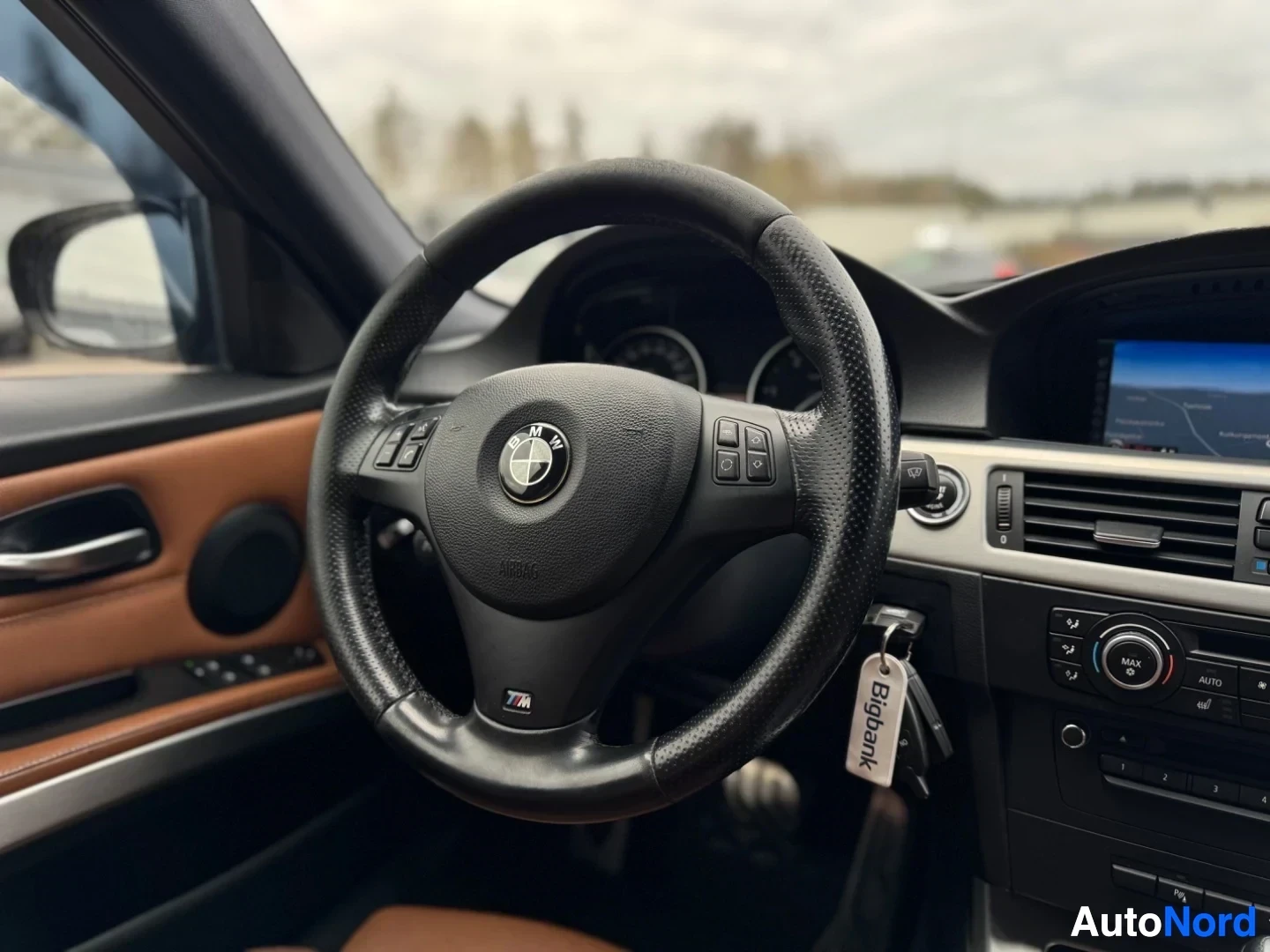Ohjaamo: Lähikuva kuljettajan puoleisesta ohjaamosta, jossa näkyy musta monitoimiohjauspyörä, jossa on BMW-logo keskellä ja "M"-logo alhaalla, sekä ruskeat nahkaiset ovi