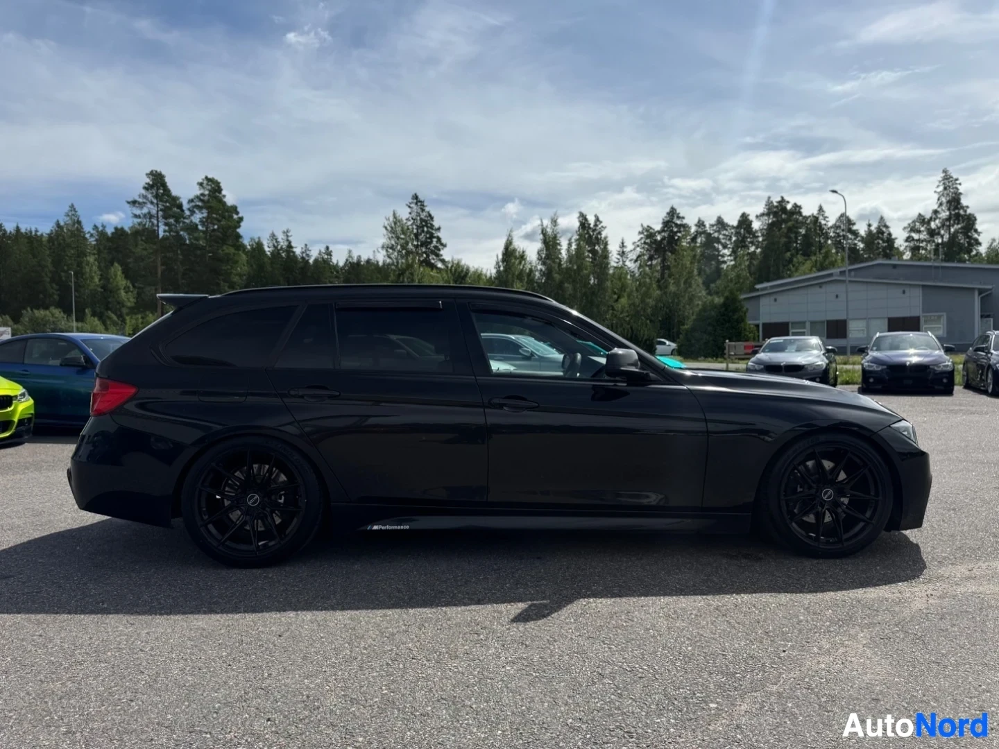 Sivulta: Musta farmariauto on pysäköity hiekkaiselle alueelle metsän ja rakennusten eteen, pilvisenä päivänä. Autossa on tummennetut takaikkunat ja mustat vanteet.