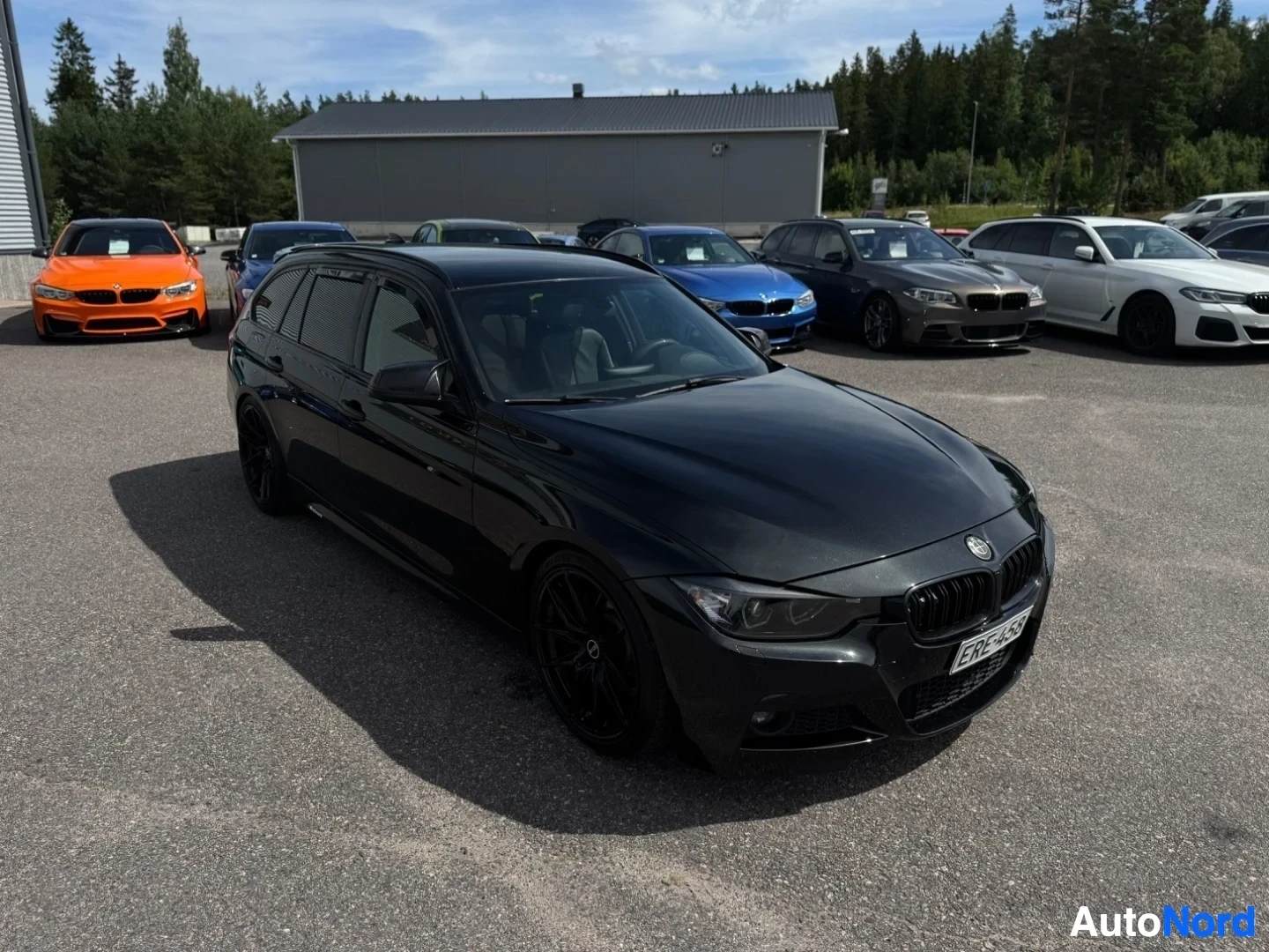 Etuviisto, nokka oikealle: Musta farmariauto pysäköitynä asfaltille, edessä oranssi auto ja taustalla useita muita autoja. Auton etuosa ja vasen kylki ovat näkyvissä. Etupuskurissa