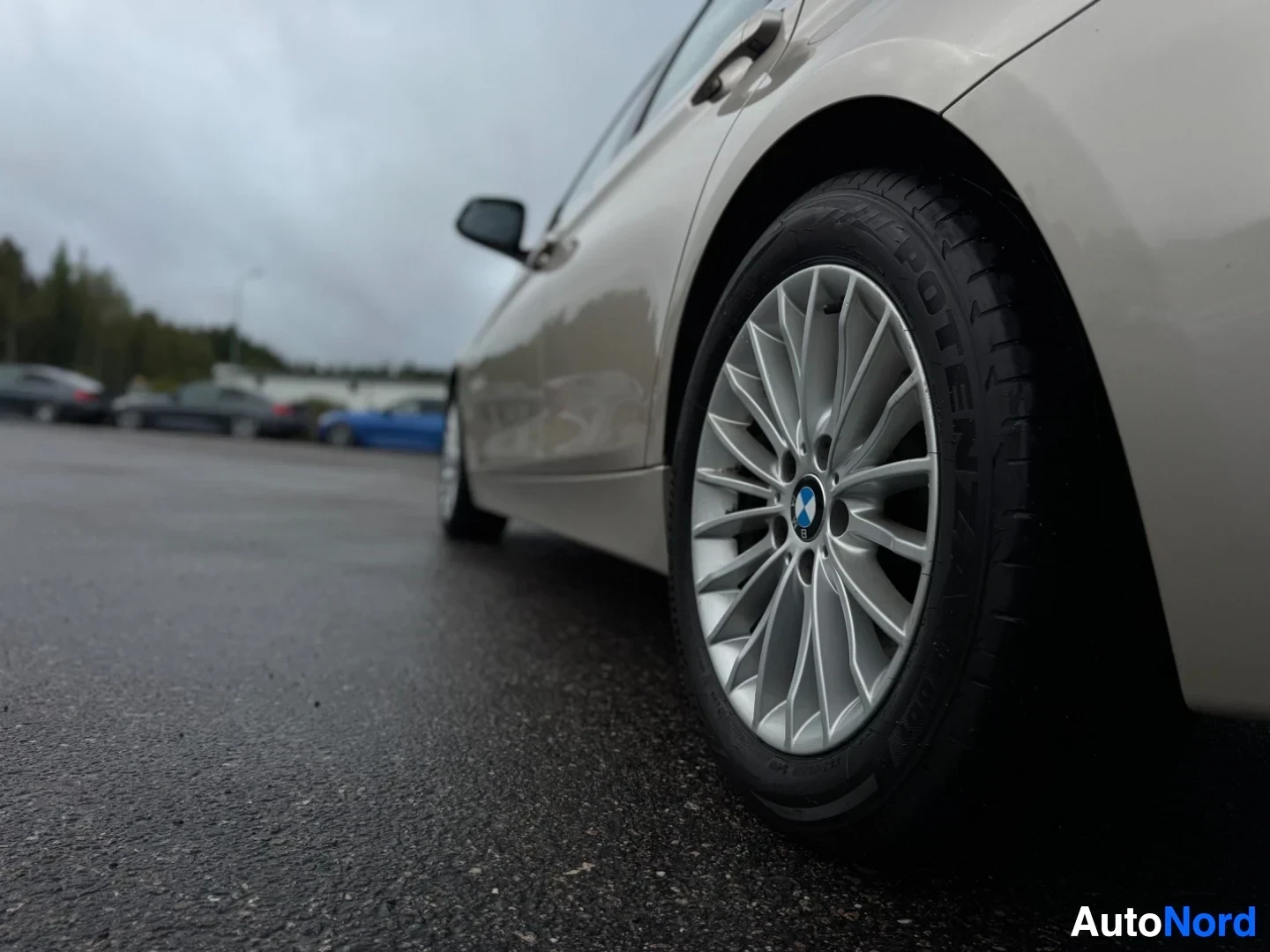 Yksityiskohta: Lähikuva auton vasemmasta etupyörästä märällä asfaltilla. Pyörässä on hopeinen monipuolavanne ja BMW-logo. Renkaan kyljessä lukee "POTENZA".