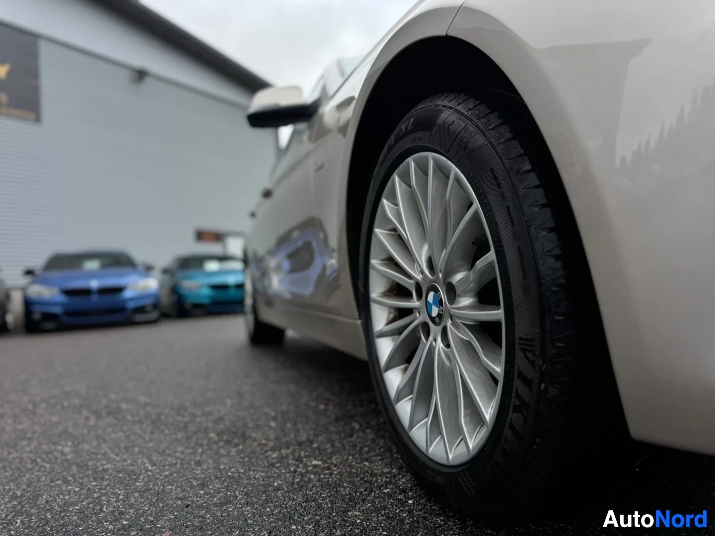 Yksityiskohta: Lähikuva auton oikeasta etupyörästä. Pyörässä on hopeinen monipuolavanne, jossa on BMW-logo. Taustalla näkyy muita autoja ja rakennus.