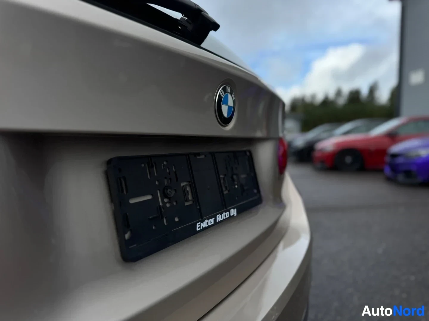 Takaviisto: Lähikuva auton takaosasta, jossa näkyy takaluukku ja tyhjä rekisterikilven kehys, jossa lukee "Enter Auto Oy". Autossa on BMW-logo, ja taustalla näkyy muita aut