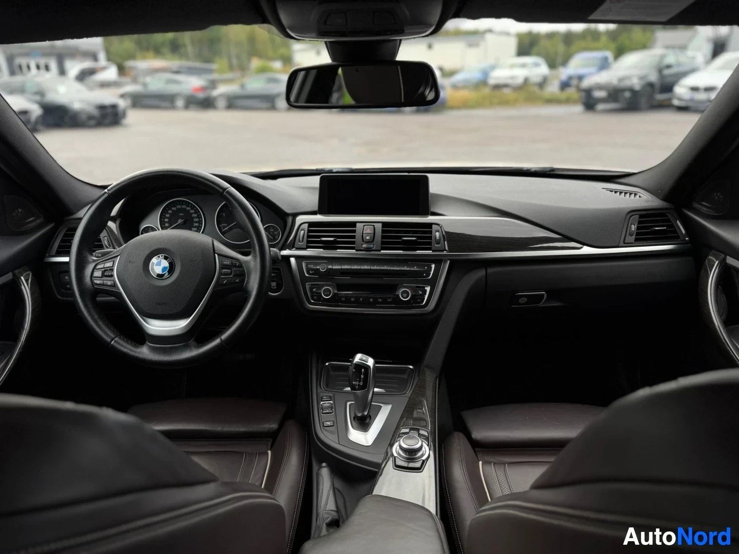 Ohjaamo: Sisäkuva auton ohjaamosta edestäpäin, keskellä ohjauspyörä ja kojelauta näytöllä. Tummat nahkaistuimet näkyvät kuvassa. Ikkunasta näkyy parkkipaikka autoineen.
