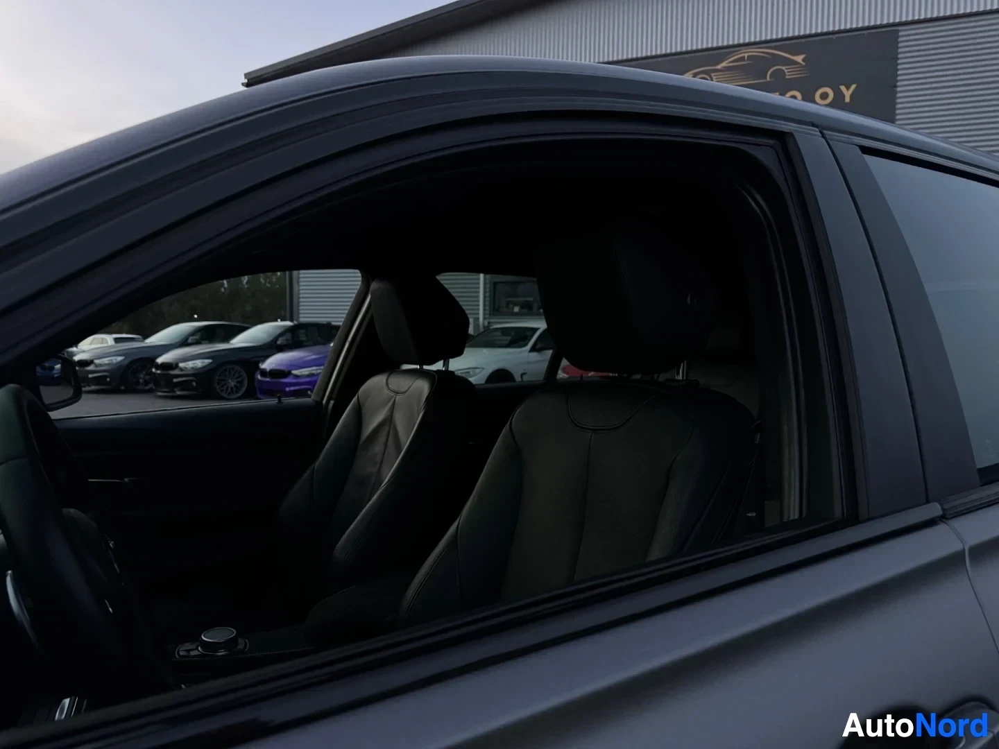 Ohjaamo: Auton matkustamon etuistuimet nahkaverhoilulla näkyvät sivuikkunan läpi, taustalla rakennus ja muita autoja.