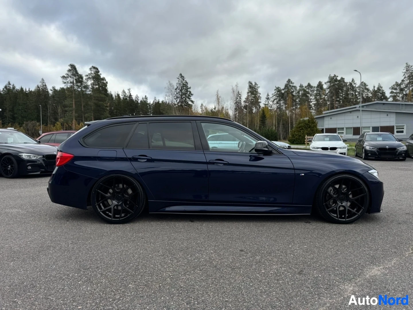 Sivulta: Tumman farmariauton vasen kylki kuvattuna sivusta. Auto on pysäköitynä ulos, ja taustalla näkyy puita ja rakennus.