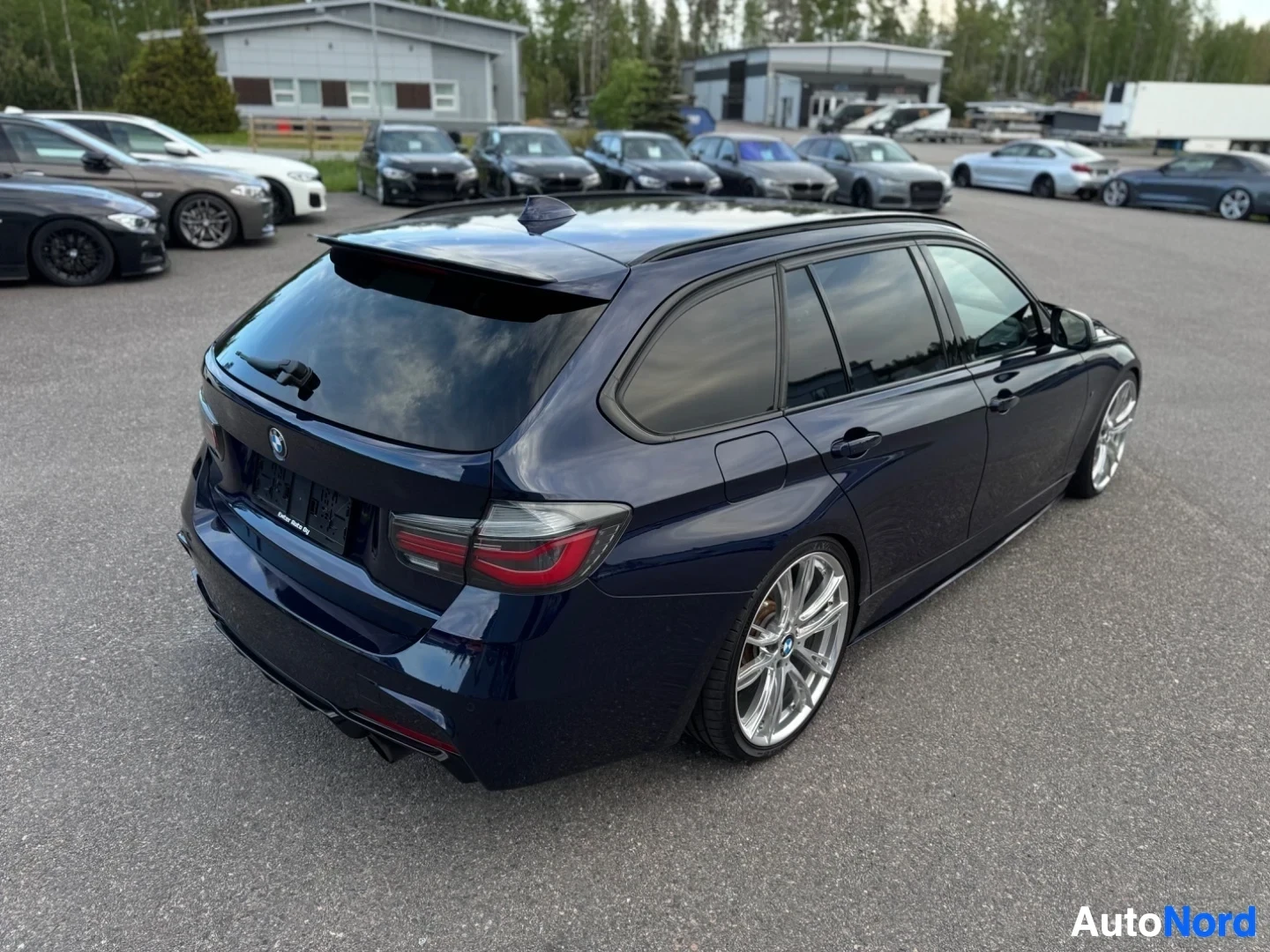 Takaviisto: Tummanpunainen BMW-farmariauto kuvattuna oikealta takaviistosta, pysäköitynä isolle asfaltoidulle alueelle muiden autojen eteen.