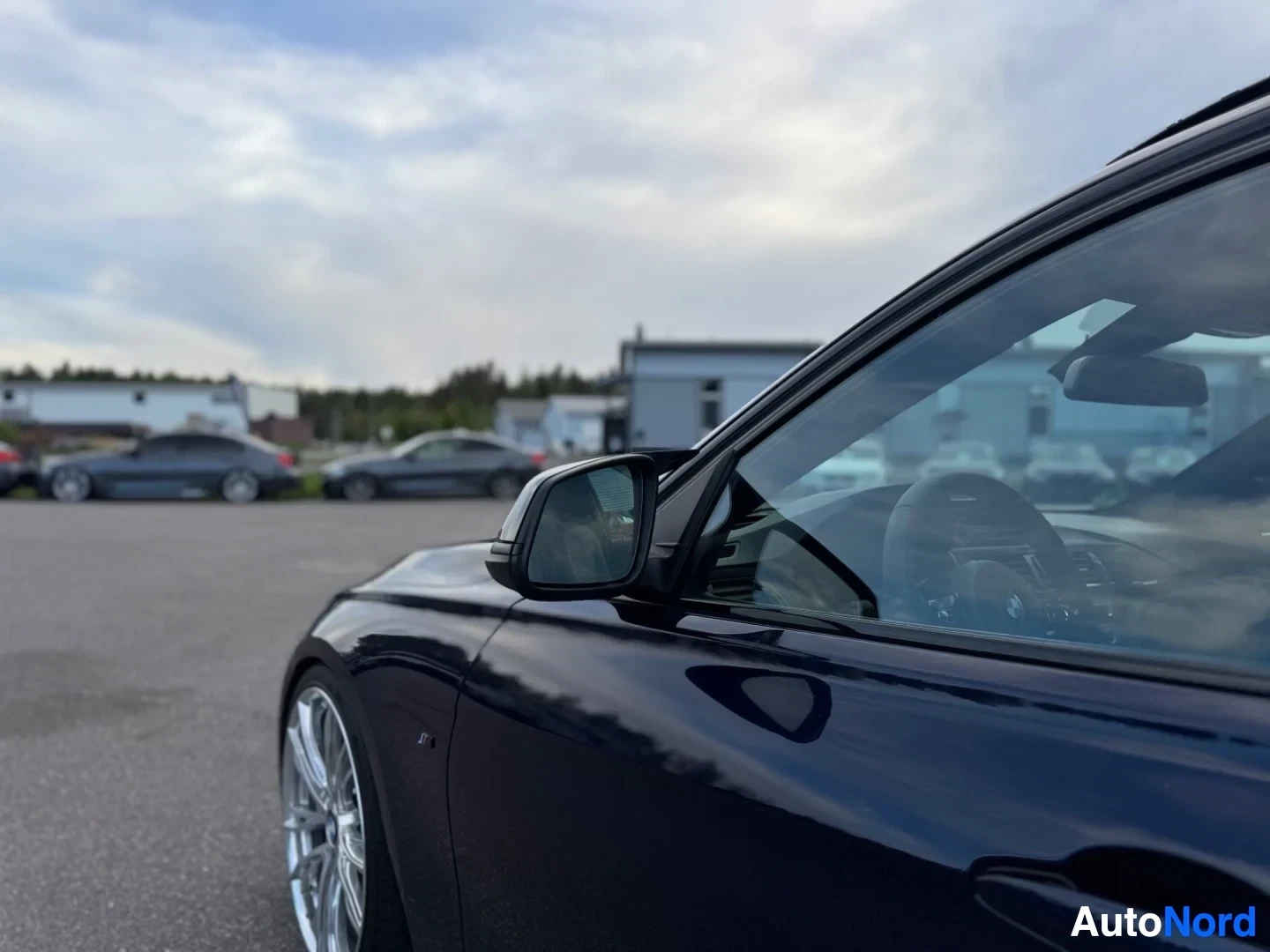 Etuviisto, nokka vasemmalle: Tummanpunainen auto, jonka keulan oikea kylki näkyy kuvassa. Auto on pysäköity asfalttialueelle, taustalla rakennuksia ja muita autoja. Kuvassa näkyy s