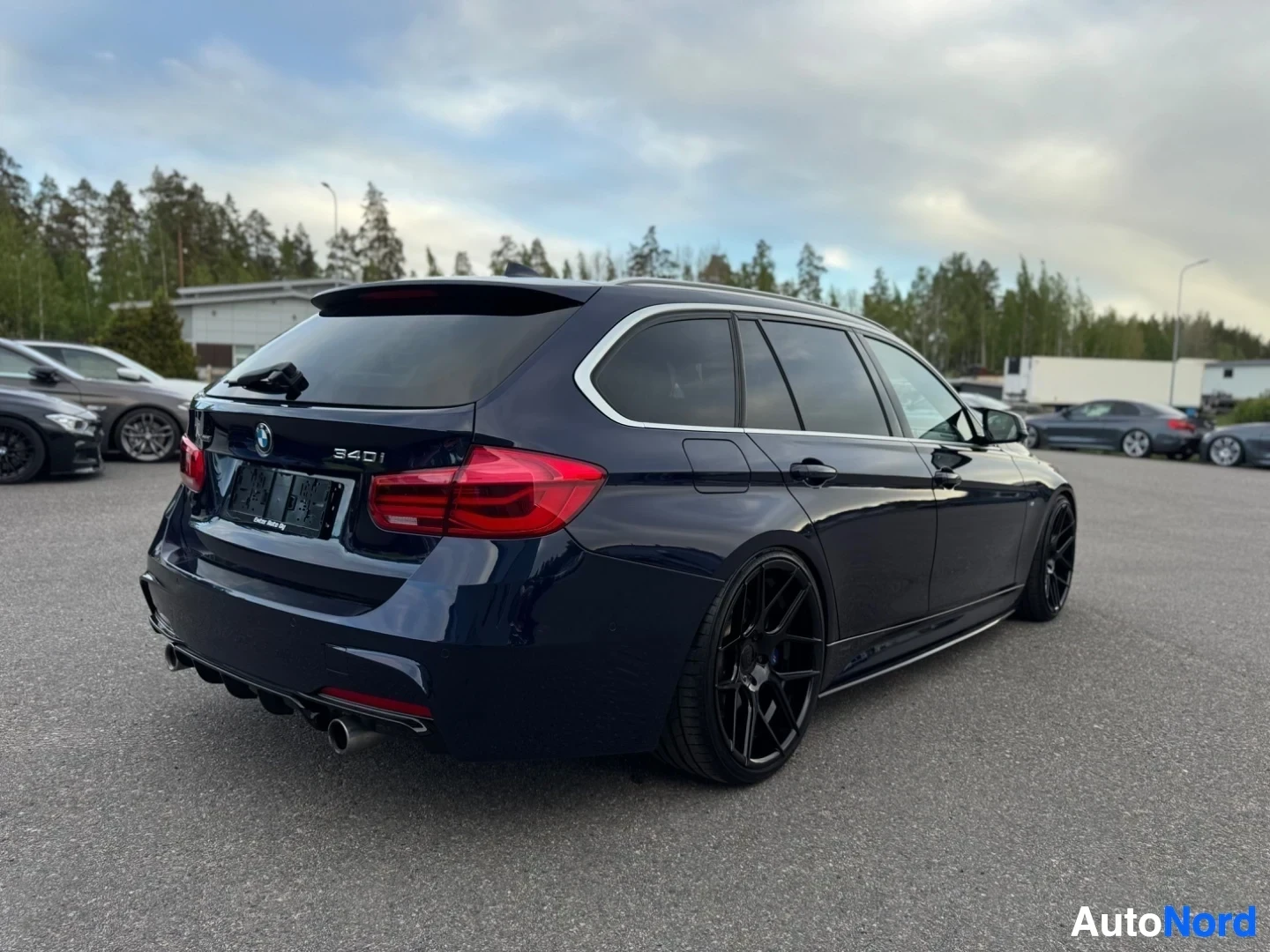 Takaviisto: Tummanpunainen BMW-farmari on pysäköity asfaltille, takaviistosta kuvattuna. Autossa on mustat monipuolaiset vanteet ja kattokaiteet. Taustalla näkyy puita ja r