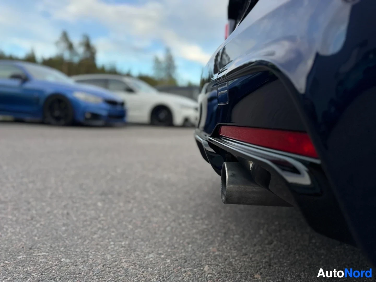 Yksityiskohta: Lähikuva auton pakoputkesta ja tummansinisestä takapuskurista, jossa on punainen heijastin. Asfaltti ja epäselvät autot näkyvät taustalla.