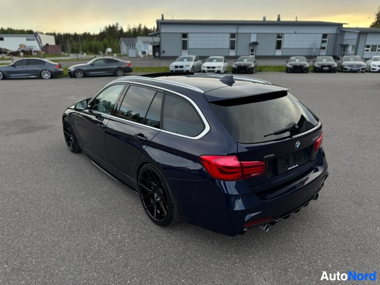 Takaviisto: Tummanpunainen BMW-farmari on pysäköity asfaltille, takaviistosta kuvattuna. Autossa on kattokaiteet ja mustat monipuolaiset vanteet. Taustalla näkyy rakennuksi