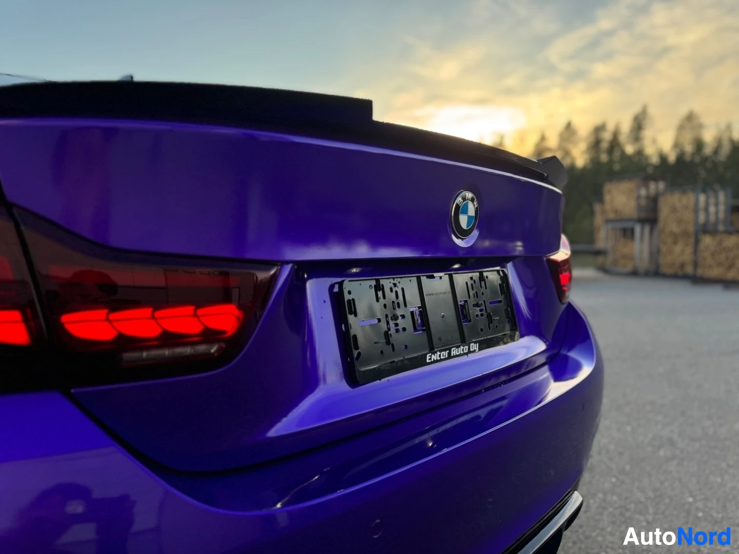 Takaviisto: Takaviisto kuva violetista autosta, jossa on musta rekisterikilven teline ja pieni spoileri takaluukussa. BMW-logo näkyy takaluukussa, ja takavalot ovat päällä.