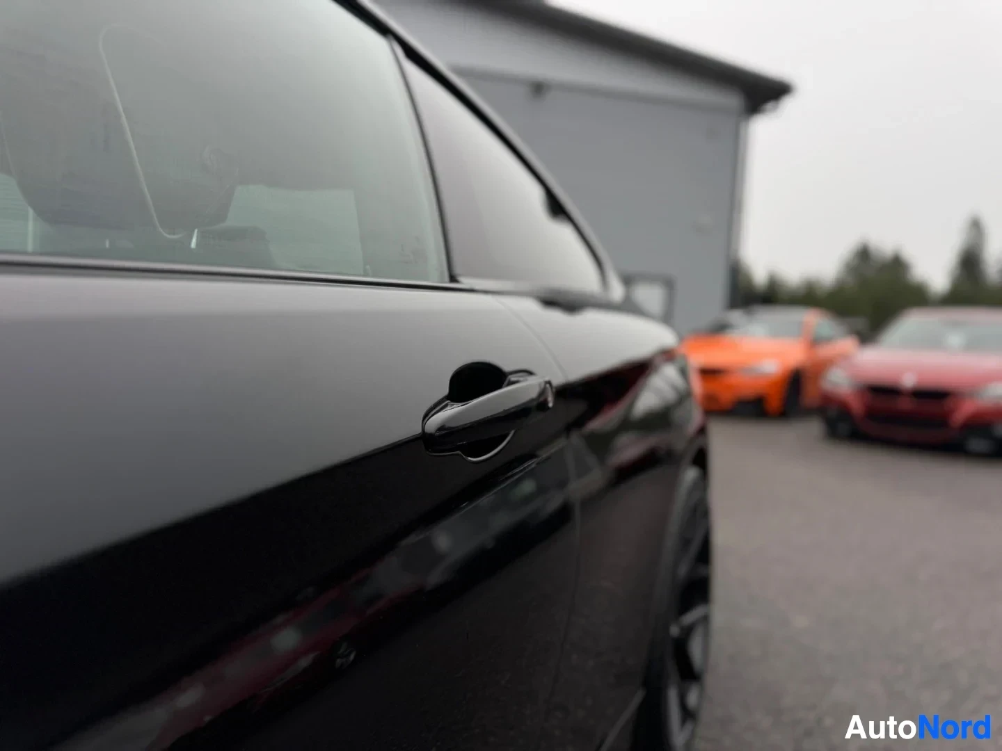 Sivulta: Mustan auton oikea kylki etuovesta taaksepäin, ulkopeilin ja ovenkahvan yksityiskohdilla. Taustalla oranssi ja punainen auto parkkipaikalla.