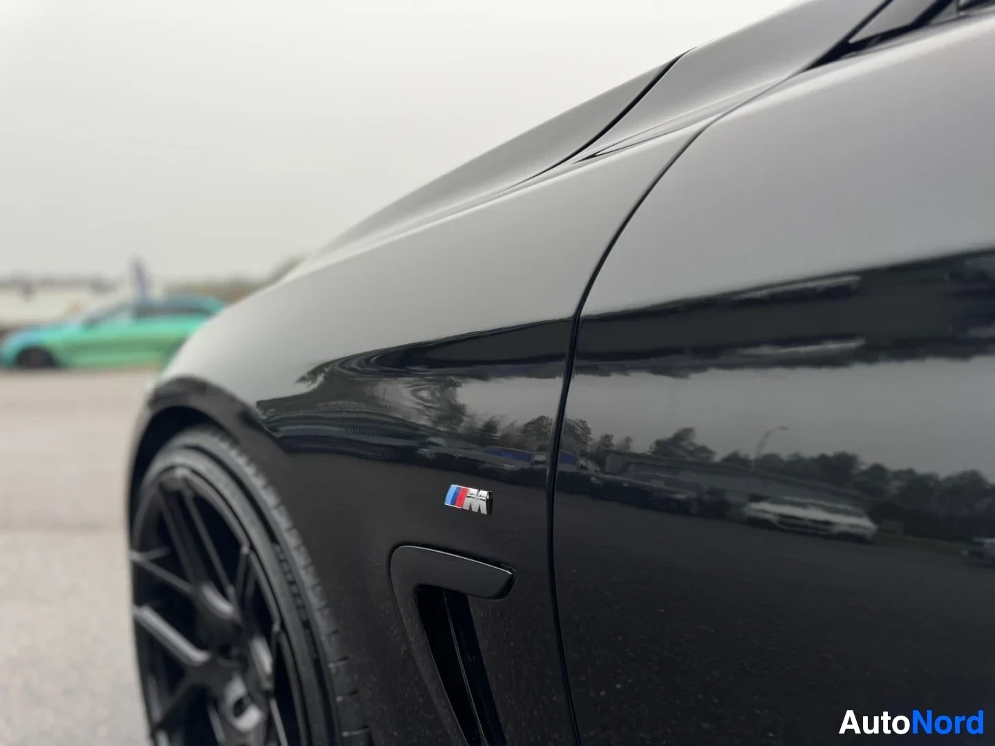 Yksityiskohta: Lähikuva mustan auton etulokasuojasta, jossa on BMW M-logo, sivuilmanvaihtoaukko ja osa mustasta kevytmetallivanteesta.