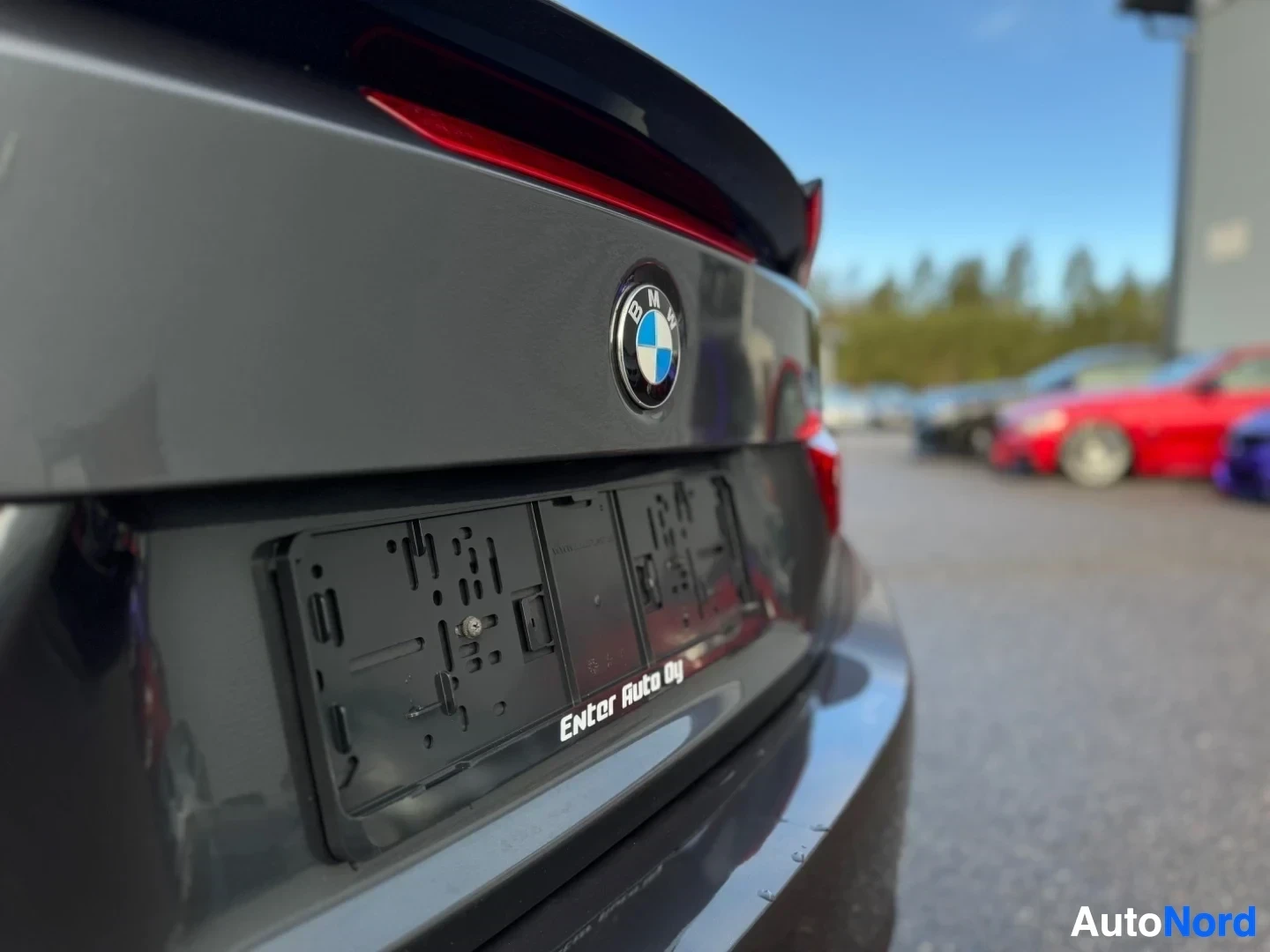 Yksityiskohta: Lähikuva auton tummanharmaan takakontin alaosasta. BMW-logo on keskellä ja sen alla on tyhjä rekisterikilven pidike, jossa lukee "Enter Auto Oy". Taustalla on p