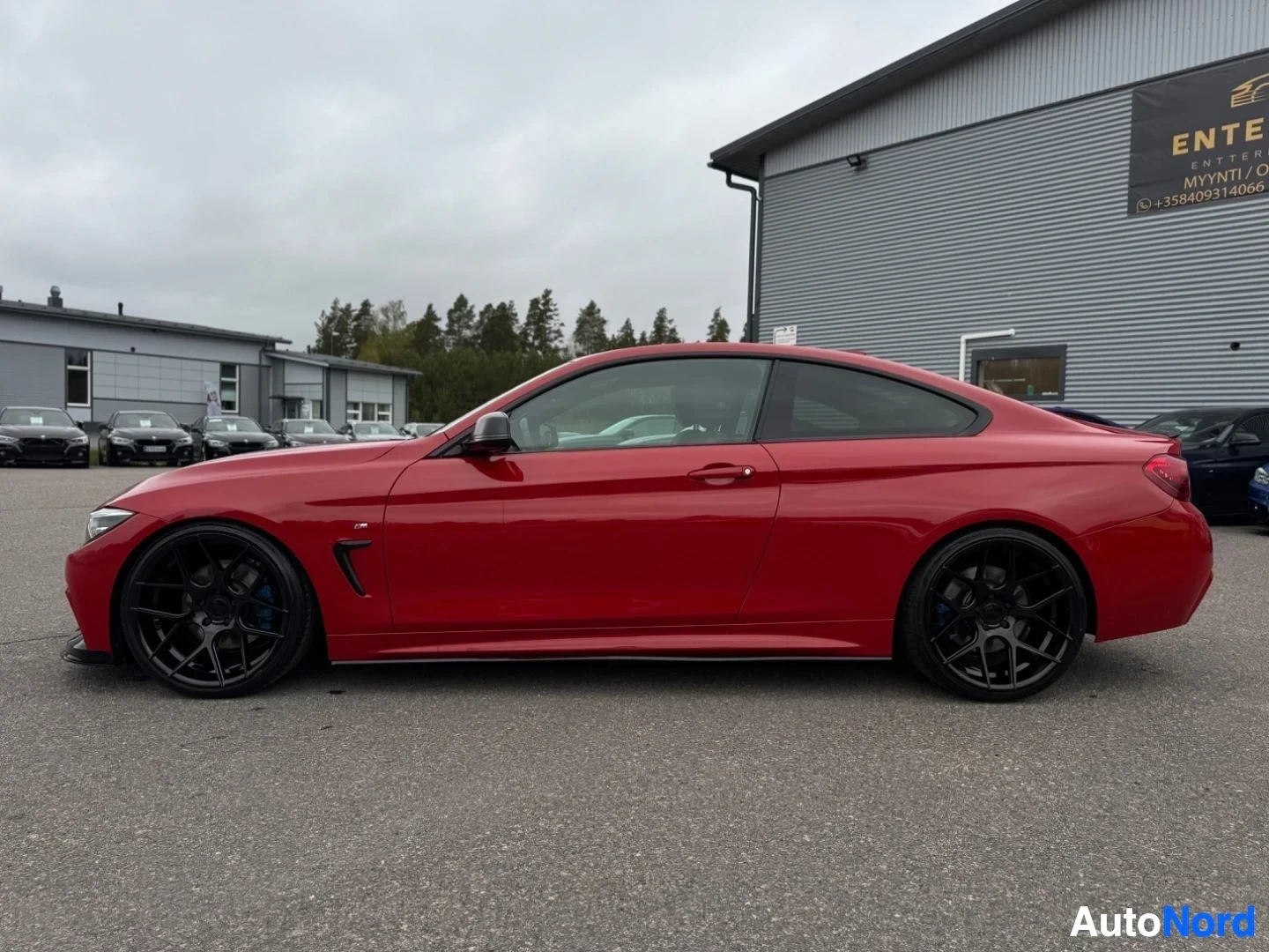 Sivulta: Punainen BMW kuvattuna suoraan kyljestä ulkona, taustalla rakennus ja puita. Autossa on mustat monipuolaiset kevytmetallivanteet.