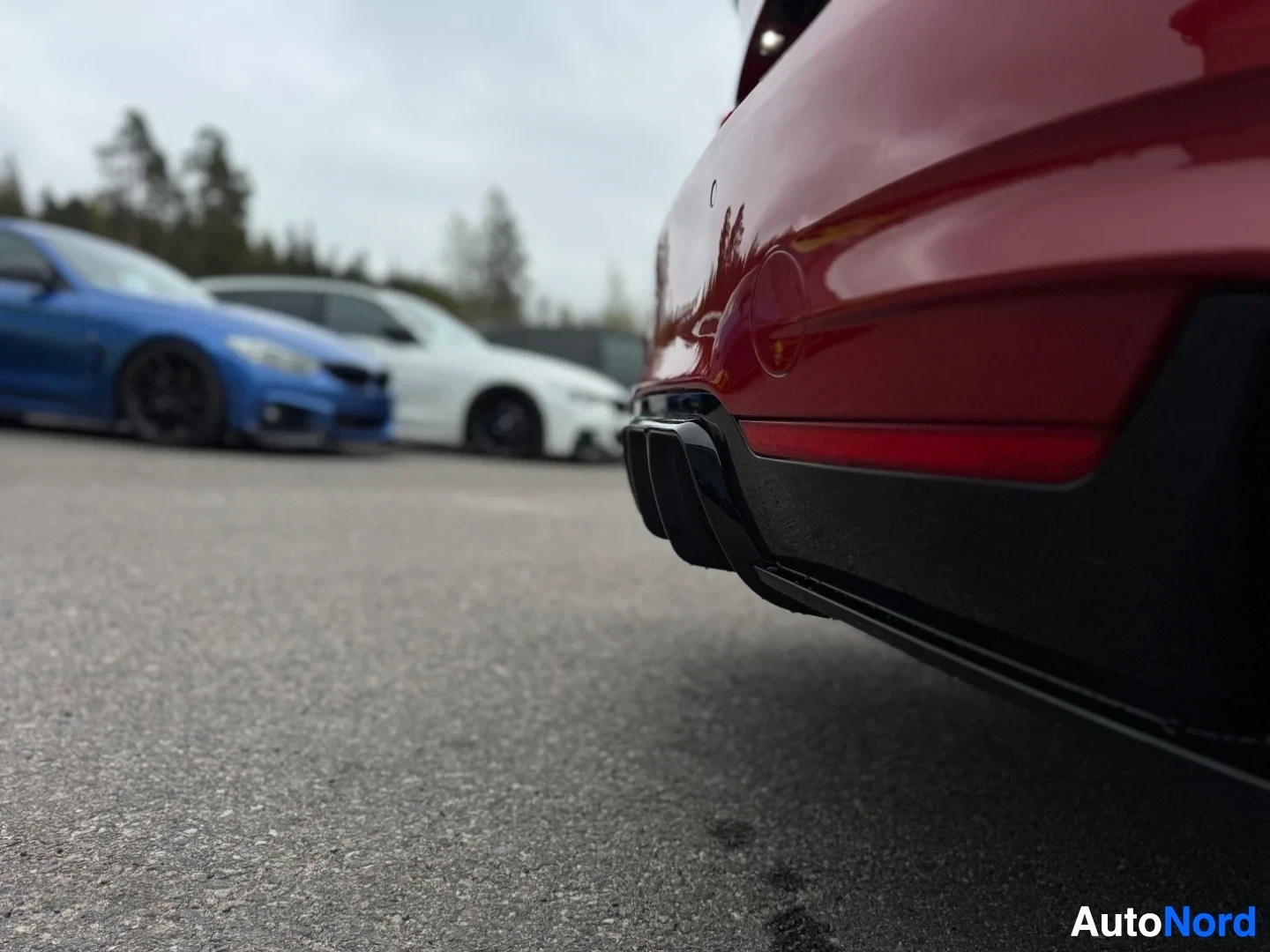 Takaviisto: Punaisen auton takaosa alhaalta kuvattuna, näkyvissä musta puskuri ja kaksi pakoputken ulostuloa. Taustalla epäselviä autoja.
