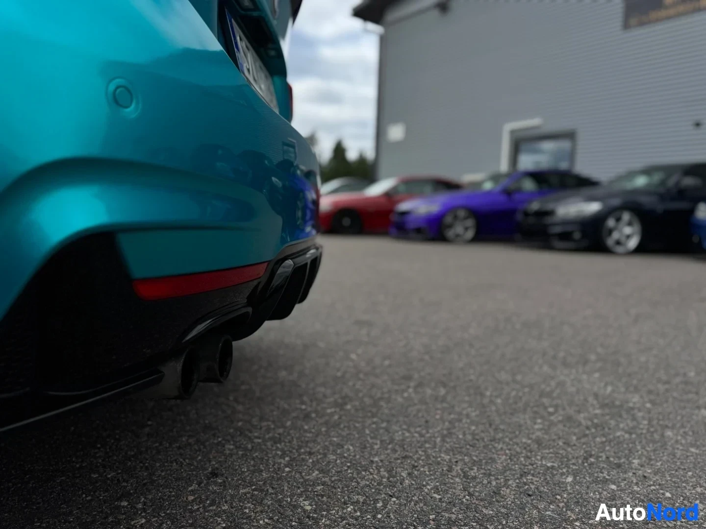 Yksityiskohta: Lähikuva turkoosin auton takapuskurista ja pakoputkista. Kaksi pakoputken ulostuloa näkyy auton oikealla puolella. Taustalla on sumeasti muita autoja.