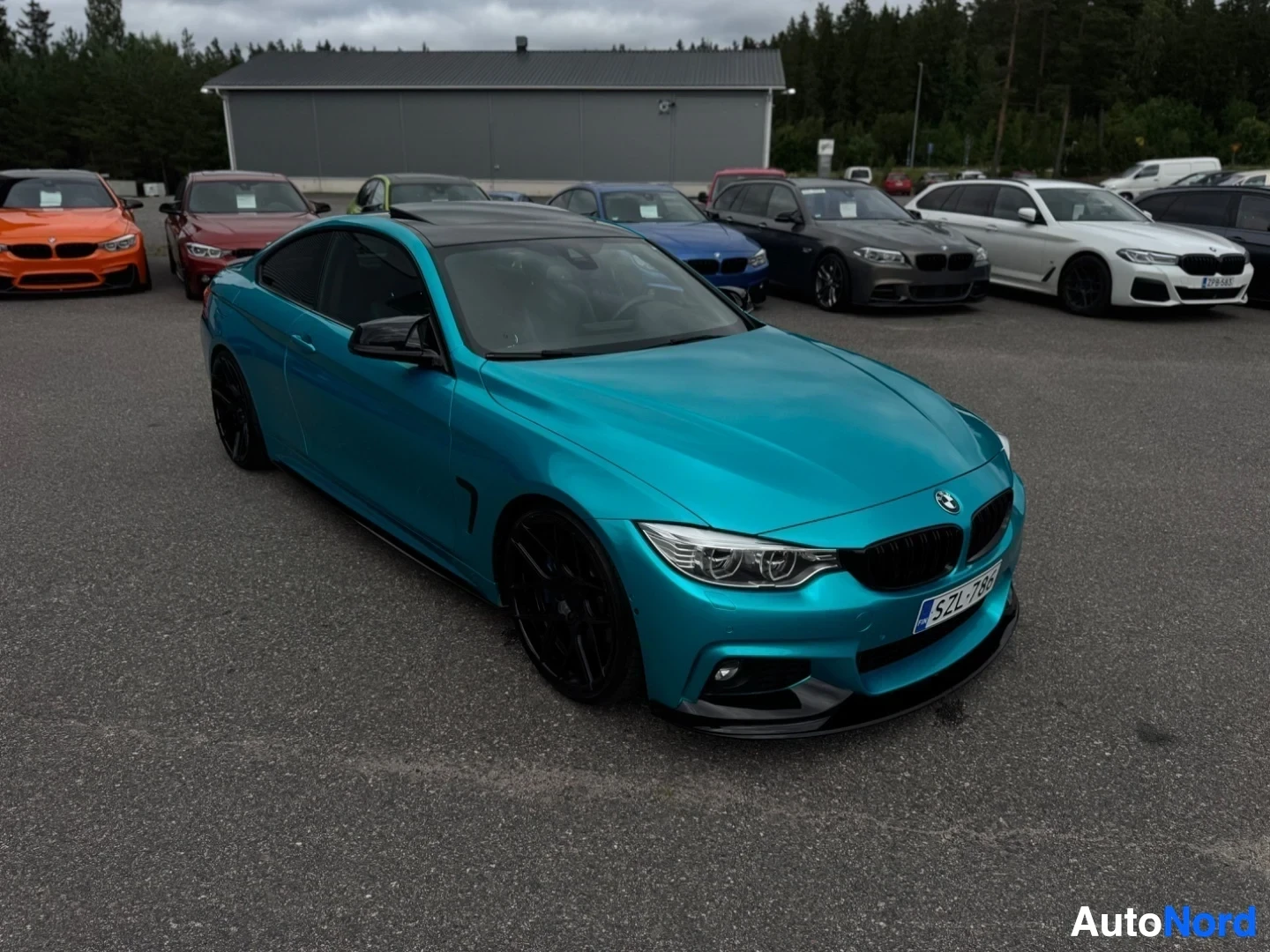 Etuviisto, nokka oikealle: Sinisenvihreä coupé-auto on pysäköity ulkona asfalttiparkkipaikalle. Etupuskurissa on musta lippa ja sumuvalot. Mustat kevytmetallivanteet näkyvät. Taust