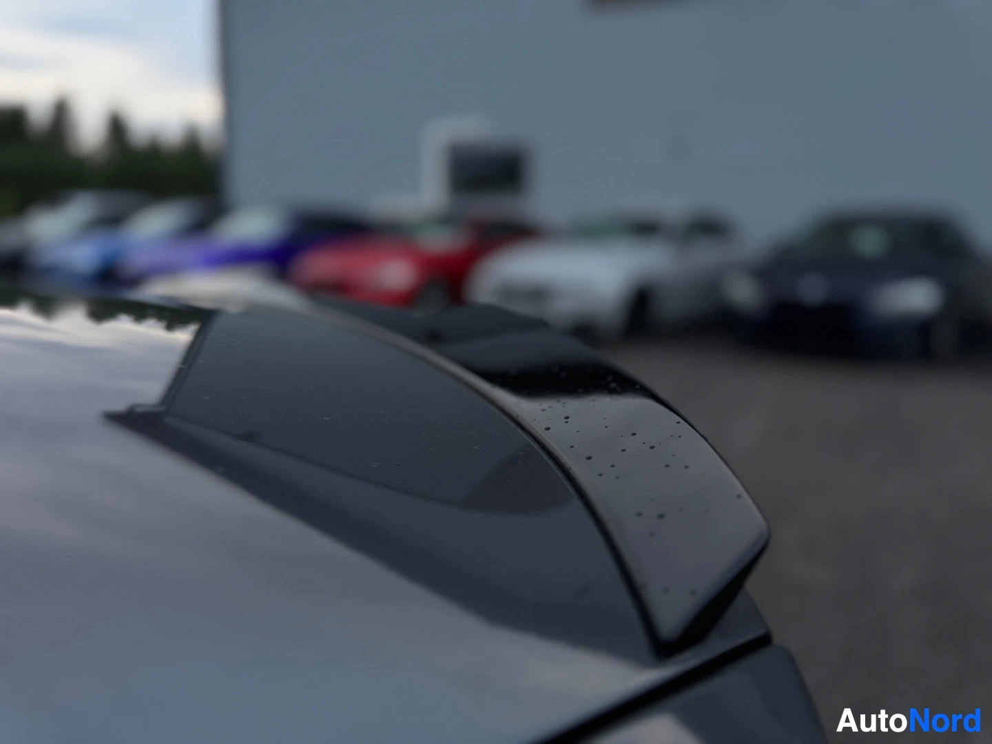 Yksityiskohta: Lähikuva auton takaluukkuun integroidusta mustasta puskurista tai spoilerin reunasta. Puskurissa on pieniä pisaroita. Taustalla useita autoja parkkipaikalla.