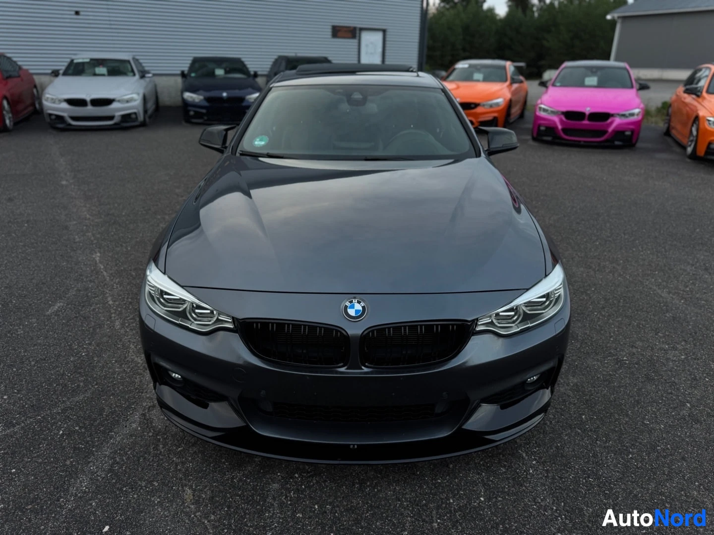 Edestä: Tummanharmaa BMW-henkilöauto edestä päin kuvattuna. Autossa on ajovalot päällä ja musta maski. Taustalla on useita muita autoja ja rakennus.