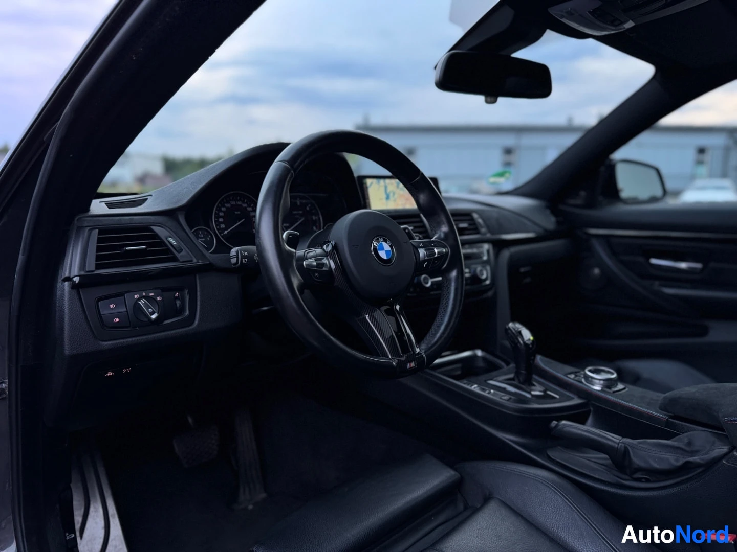Ohjaamo: Kuva auton ohjaamosta. Nahkaverhoiltu monitoimiohjauspyörä BMW-logolla ja keskikonsoli vaihteenvalitsimineen on näkyvissä. Kojelaudassa on näyttö.