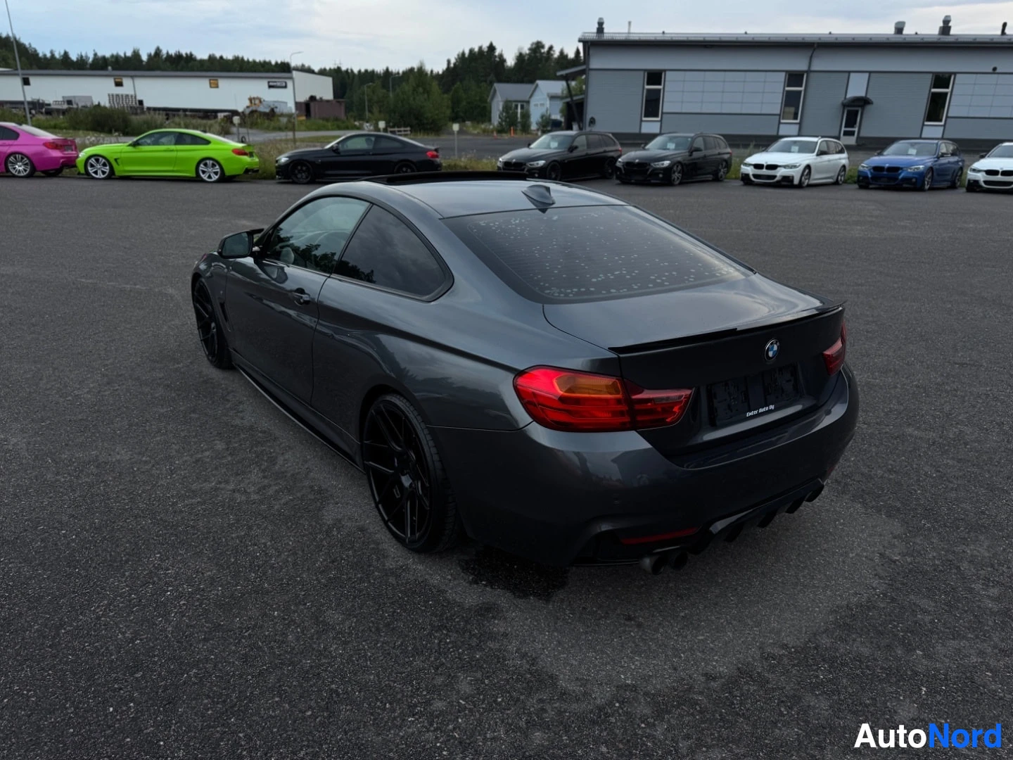 Takaviisto: Tummanharmaa BMW-henkilöauto, jossa on tummat kevytmetallivanteet ja takaspoileri, pysäköitynä asfaltille ulkona. Takavalot ja oikea kylki näkyvät. Auto on kuva