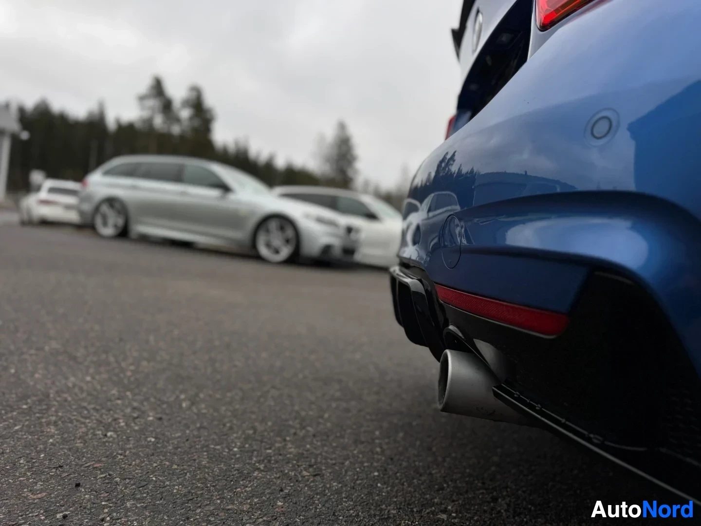 Takaviisto: Osittainen takakuva sinisestä autosta, joka on pysäköitynä päällystetylle alueelle. Pakoputken pää ja takapuskuri näkyvät oikealla alhaalla. Taustalla näkyy mui