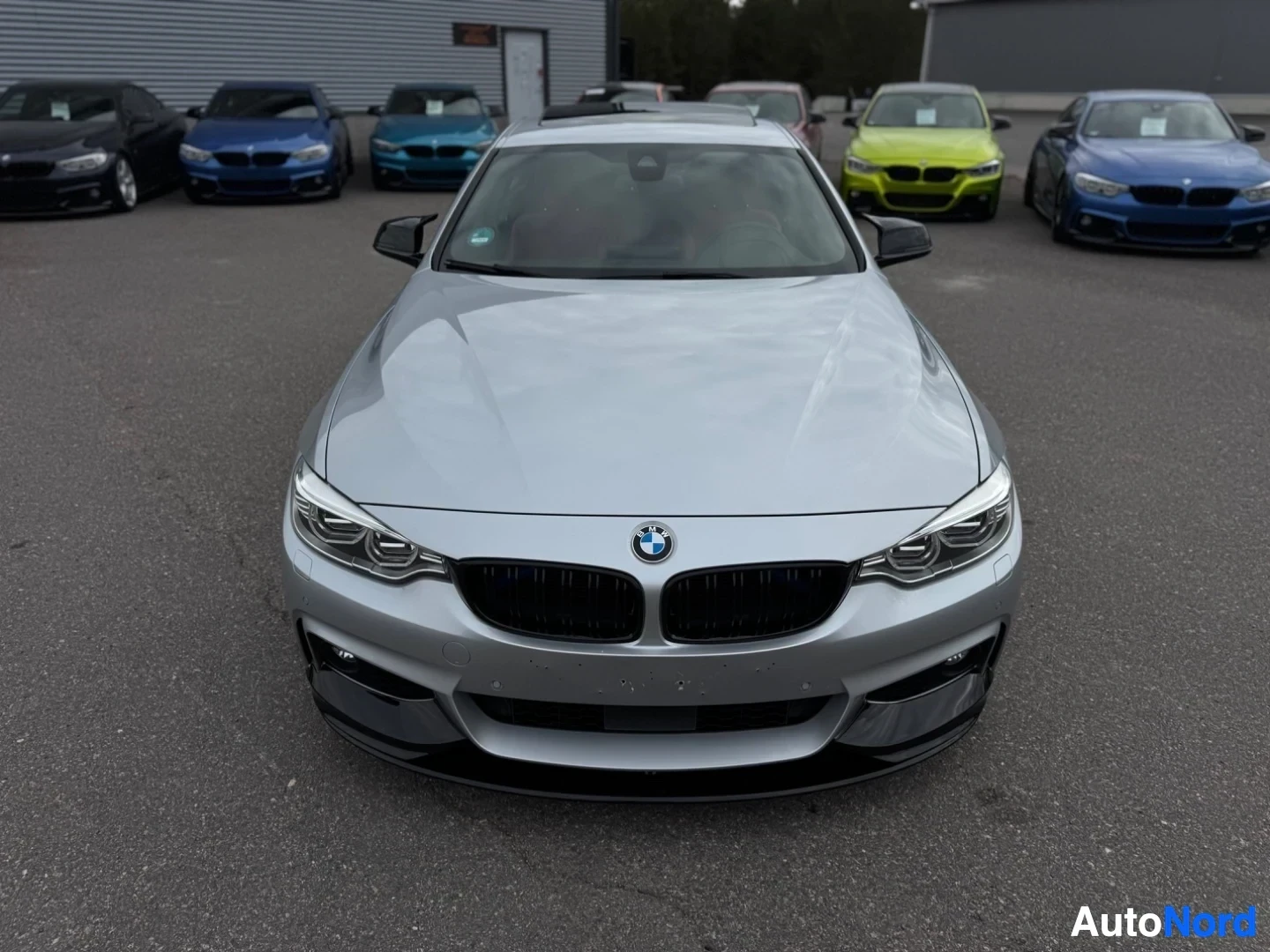 Edestä: Hopeanvärinen BMW pysäköitynä rakennuksen eteen muiden autojen kanssa. Kuva on otettu suoraan edestä, näyttäen auton keulan.