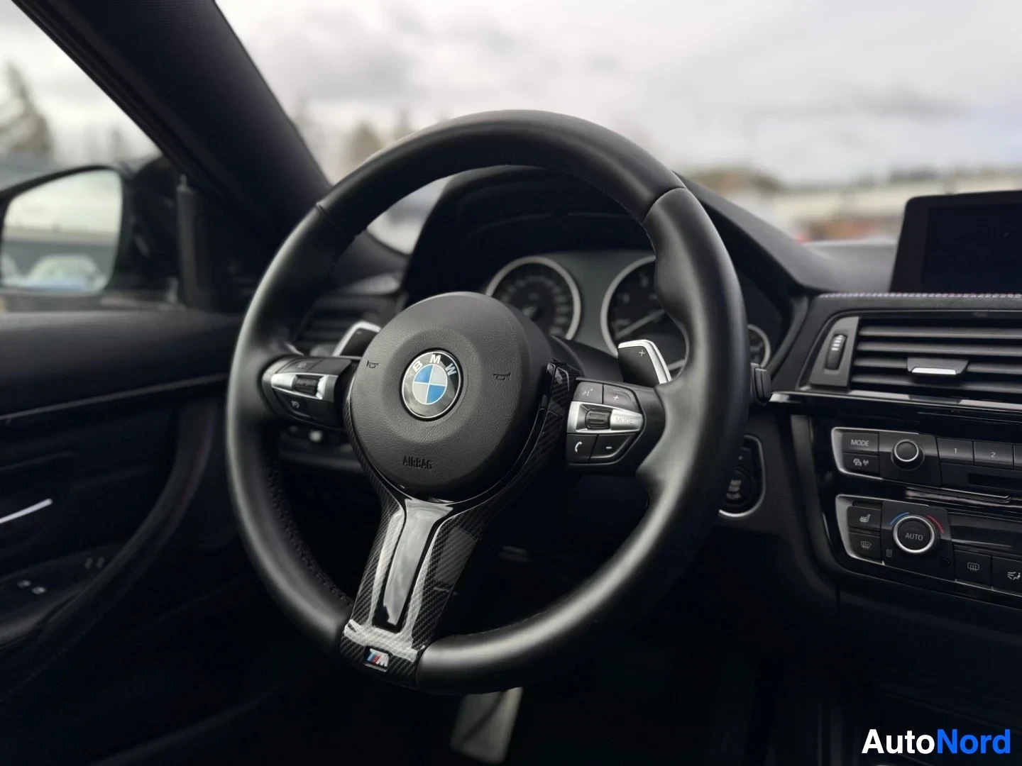 Yksityiskohta: Lähikuva kolmipuolaisesta ohjauspyörästä, jossa on BMW-logo ja 'AIRBAG'-merkintä. Ohjauspyörässä on painikkeita ja hiilikuitua jäljittelevä koriste.