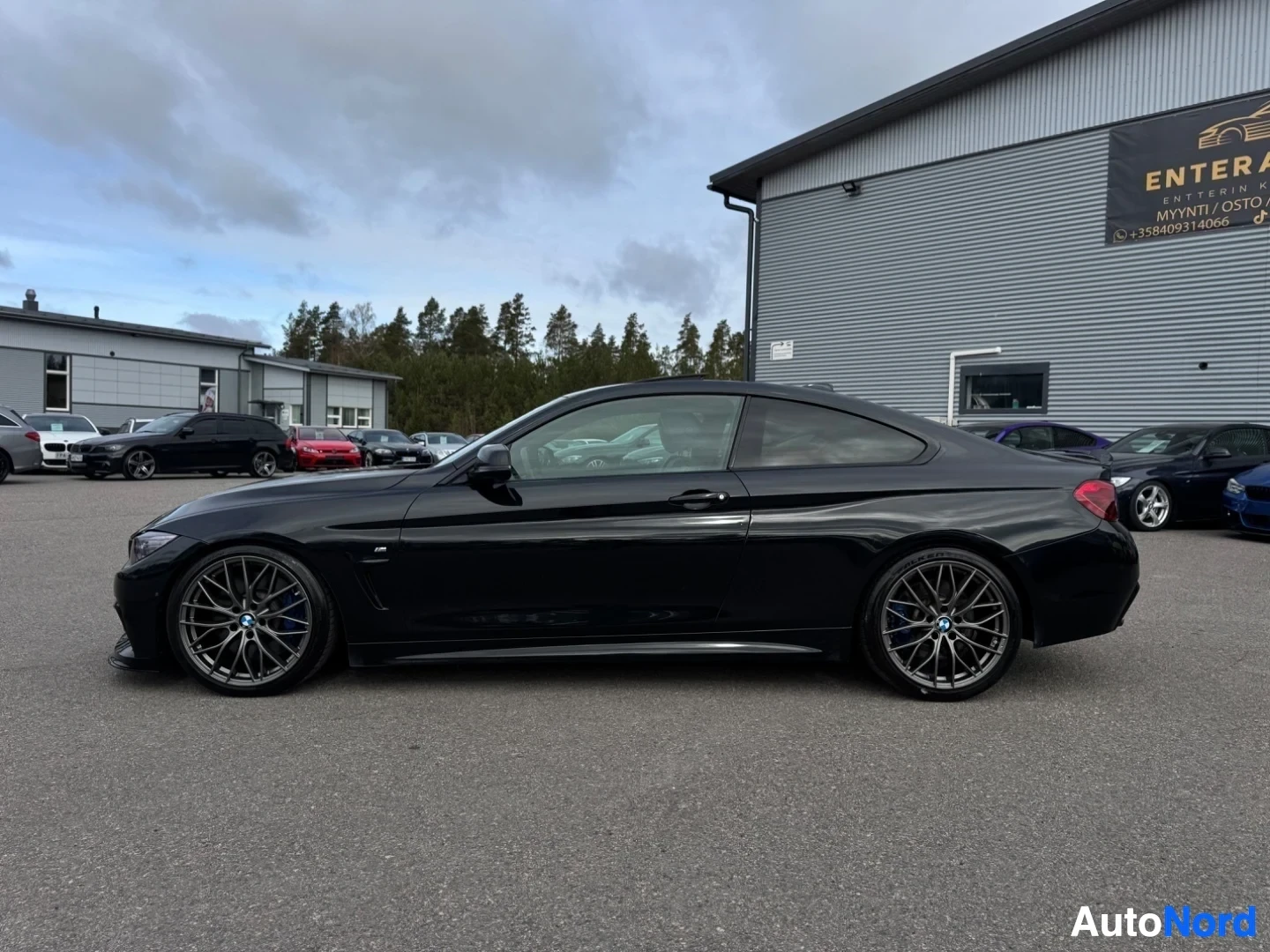 Sivulta: Musta coupé parkkipaikalla, oikealla autoliikkeen mainostaulu. Takavasemmalta kuvattu. Ikkunoissa heijastuksia.