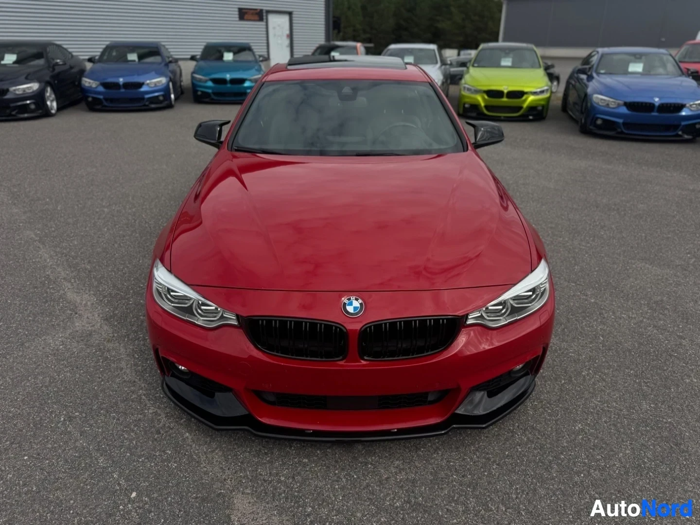 Edestä: Punainen BMW-henkilöauto on kuvattu edestäpäin. Autossa on musta etusäleikkö ja mustat yksityiskohdat etupuskurissa. Ajovalot ovat päällä. Taustalla näkyy useit