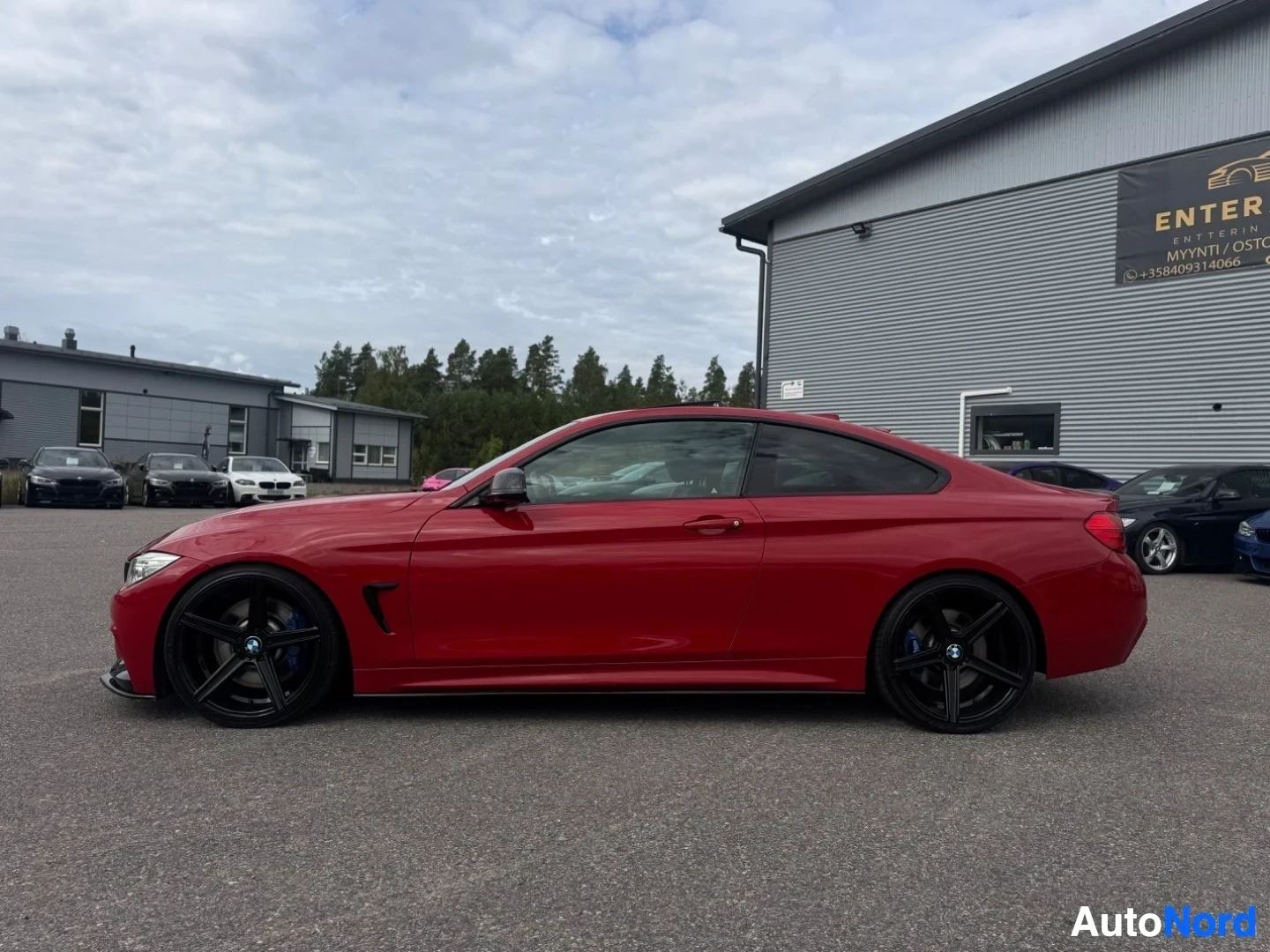 Sivulta: Punainen BMW 4-sarjan coupé, jossa on mustat monipuolaiset kevytmetallivanteet ja lasinen kattoluukku, pysäköitynä ulkona autokaupan pihalle. Auton oikea kylki