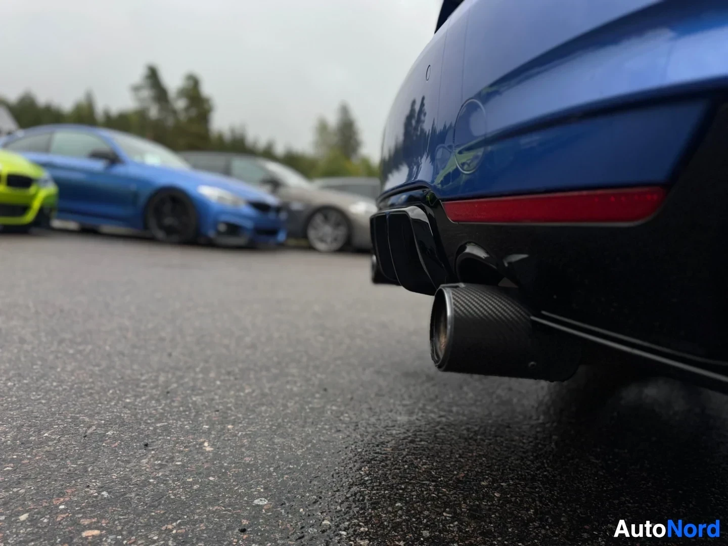 Yksityiskohta: Lähikuva sinisen auton takaosasta, jossa näkyy hiilikuitupintainen pakoputken ulostulo ja musta diffuusori märkällä asvaltilla. Taustalla epätarkkoja autoja.