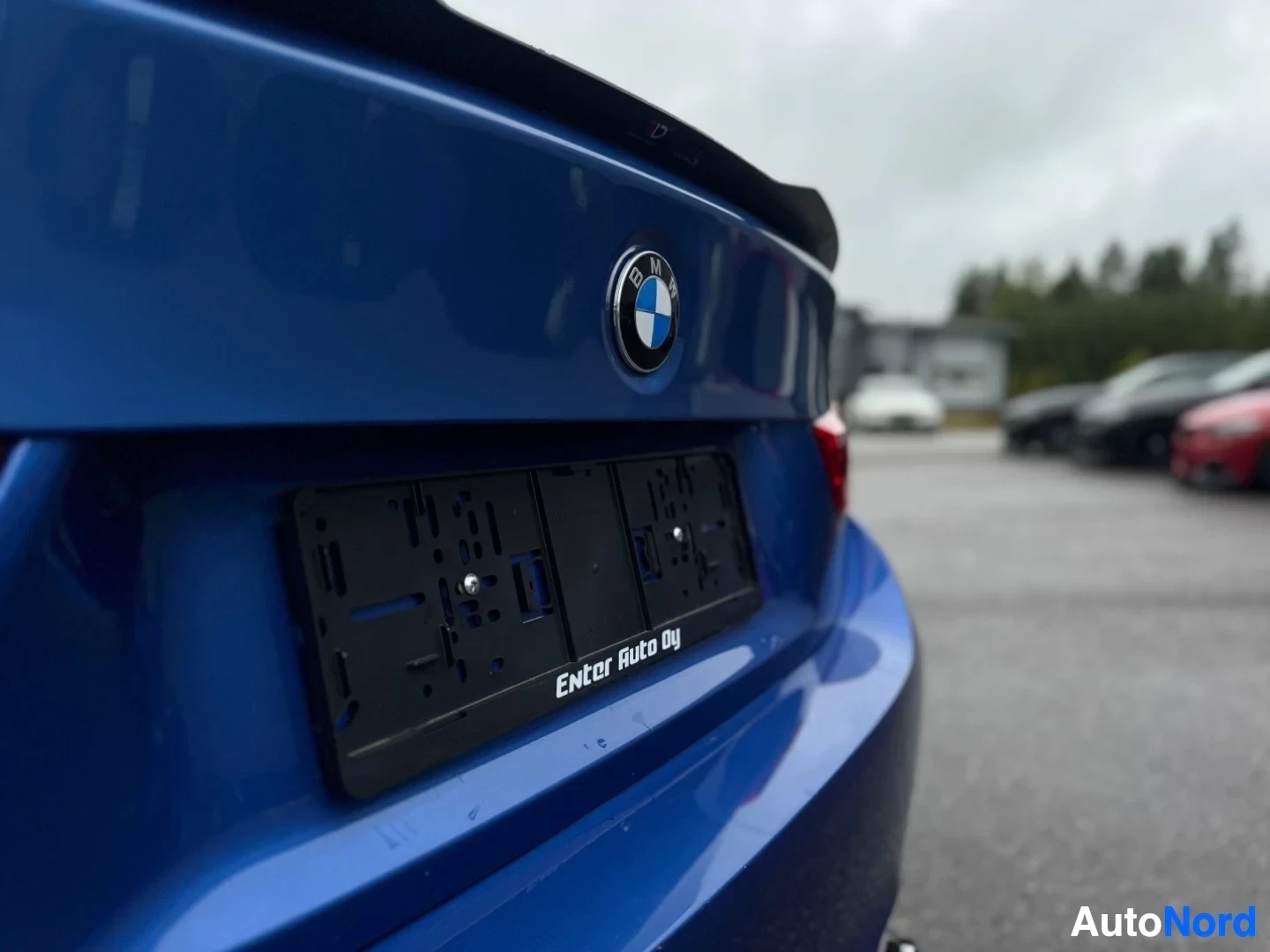Takaviisto: Lähikuva sinisen auton peräluukun oikeasta puolesta, jossa näkyy BMW-logo ja musta rekisterikilven pidike, jossa lukee "Enter Auto Oy".