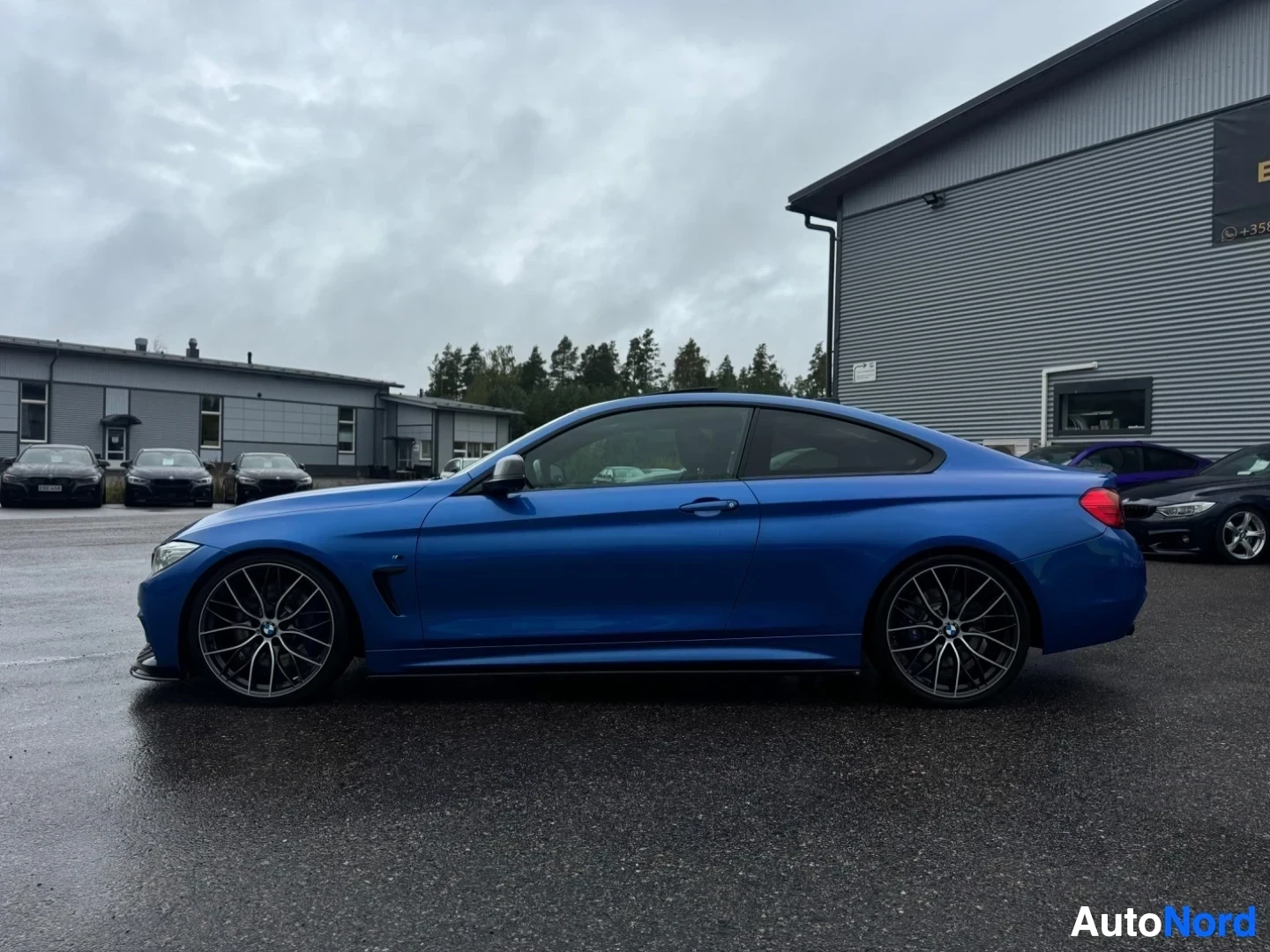 Sivulta: Sinisen BMW 4-sarjan coupen vasen kylki pysäköitynä harmaalle asvaltille rakennuksen ja puiden eteen. Autossa on mustat vanteet.