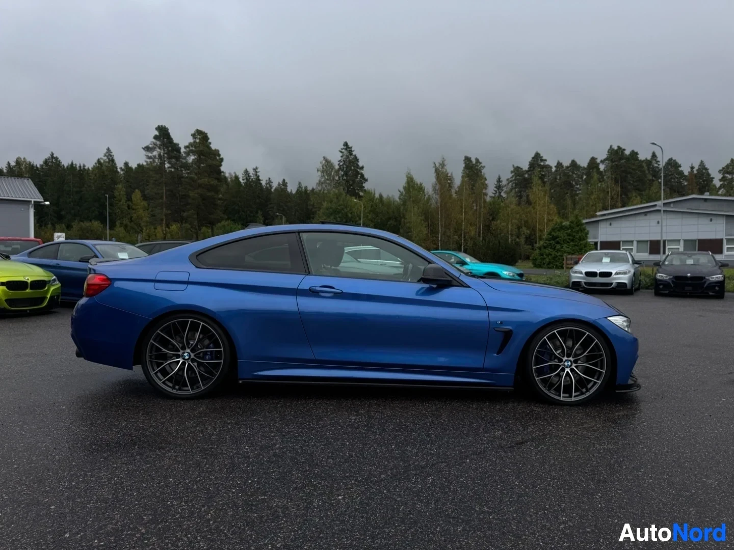 Sivulta: Sinisen BMW 4-sarjan coupen oikea kylki pysäköitynä harmaalle asvaltille, puiden ja muiden autojen eteen. Autossa on mustat vanteet.