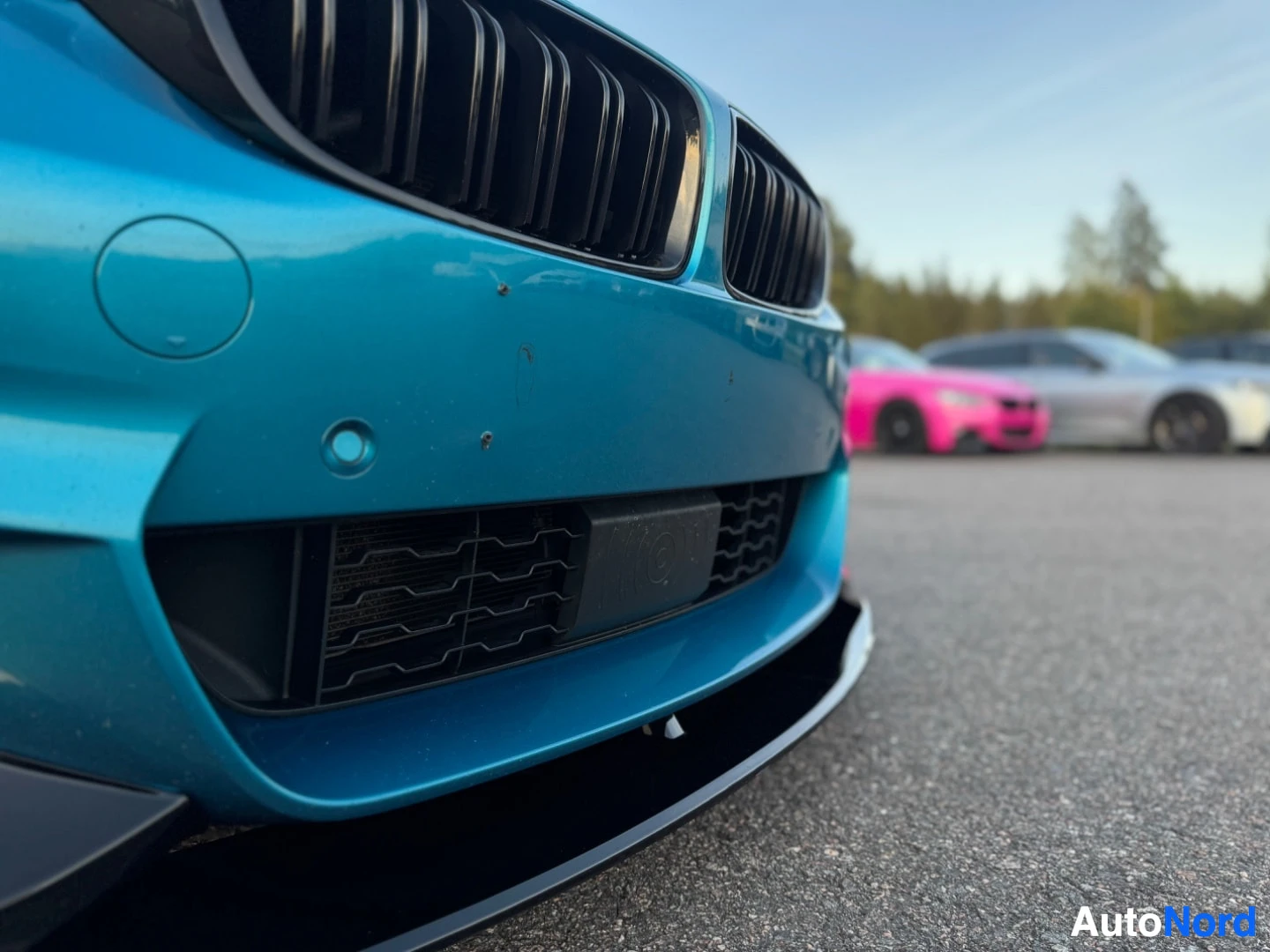 Yksityiskohta: Lähikuva turkoosin auton etuosasta, jossa näkyy musta säleikkö, alaosa ja etupuskuri antureineen. Auto on pysäköity ulos, ja taustalla on muita autoja.