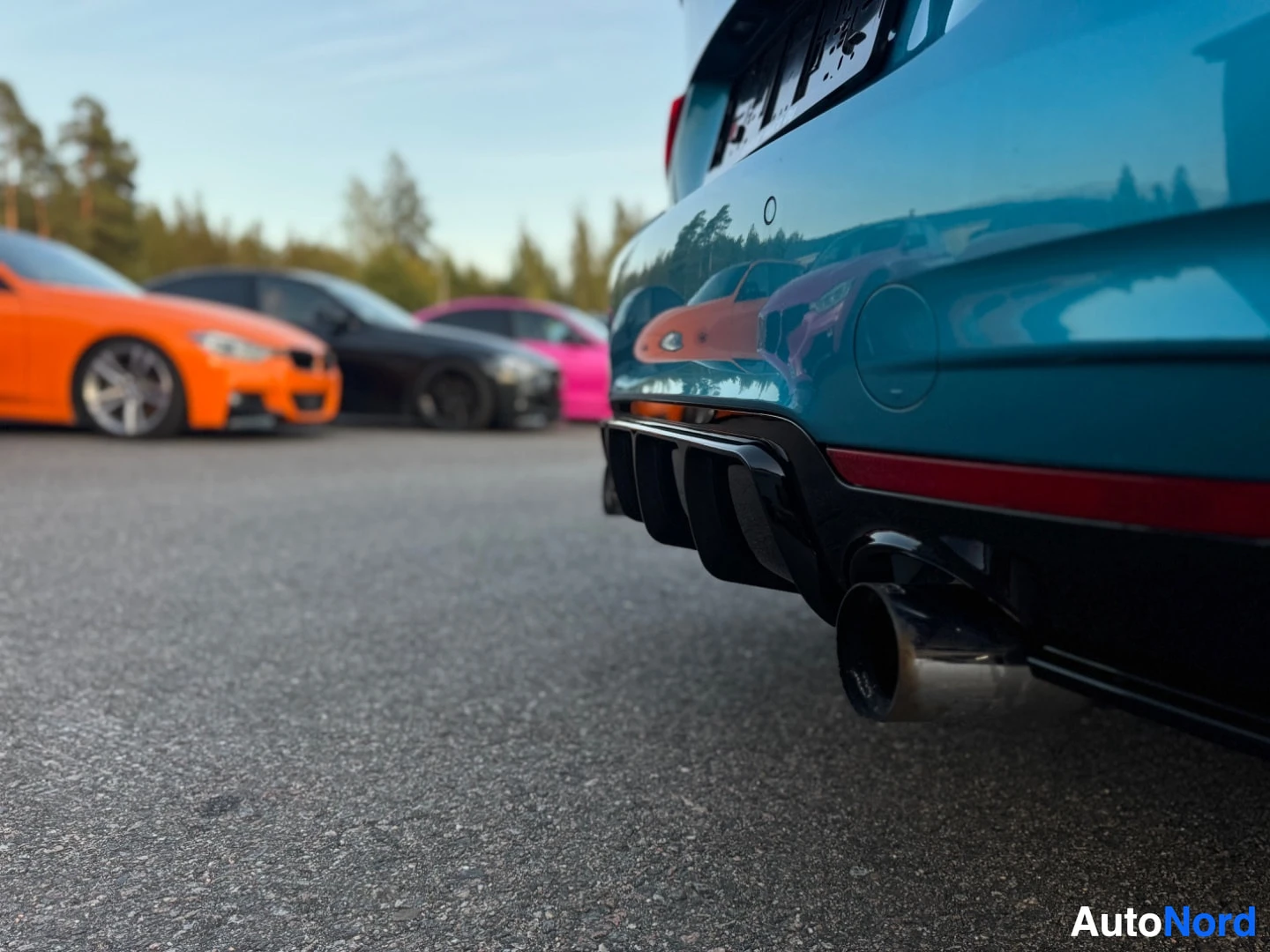 Takaviisto: Sinisen auton oikeanpuoleinen takaosa, jossa näkyy pakoputki ja diffuusori. Taustalla on rivi muita autoja, joista yksi oranssi on lähimpänä.