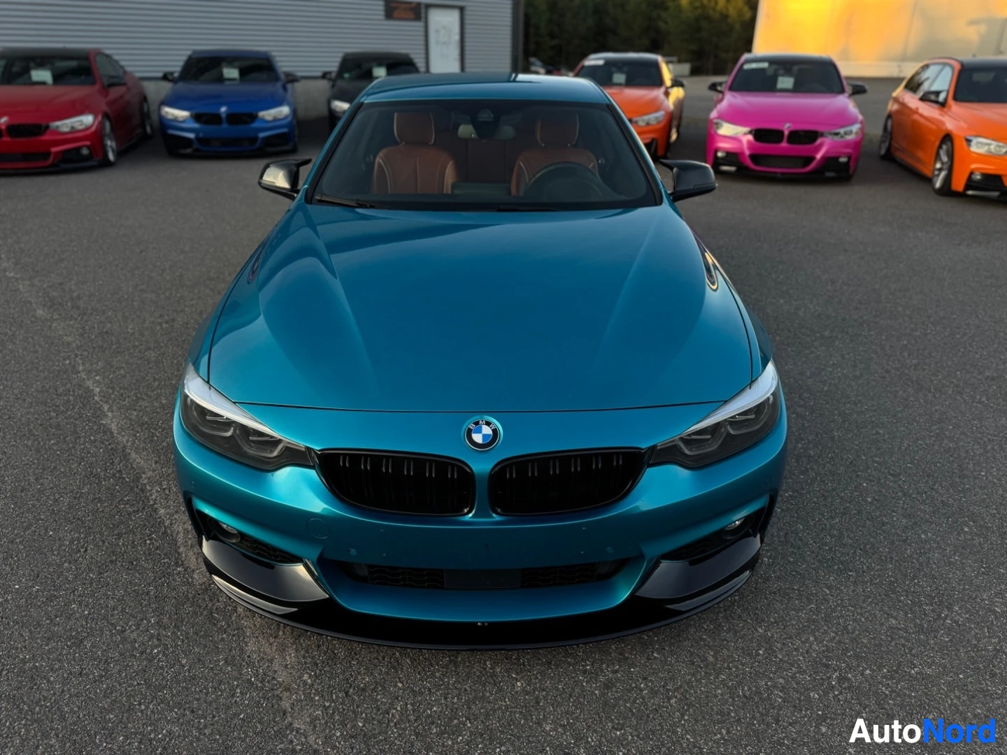 Edestä: Sinisenturkoosi BMW-henkilöauto, jossa on musta etusplitteri ja musta munuaissäleikkö, kuvattuna edestä rakennuksen edessä. Auton sisätilojen oranssinruskeat is