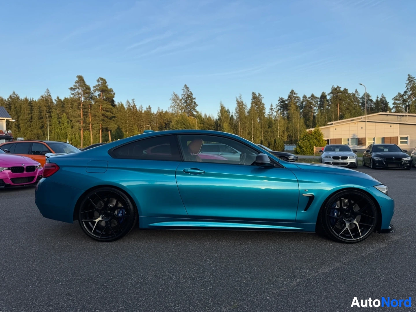Sivulta: Sininen coupé-tyyppinen auto, jonka oikea kylki on kuvattu sivuprofiilista. Auto on pysäköity ulos, ja taustalla näkyy puita ja kirkas taivas.