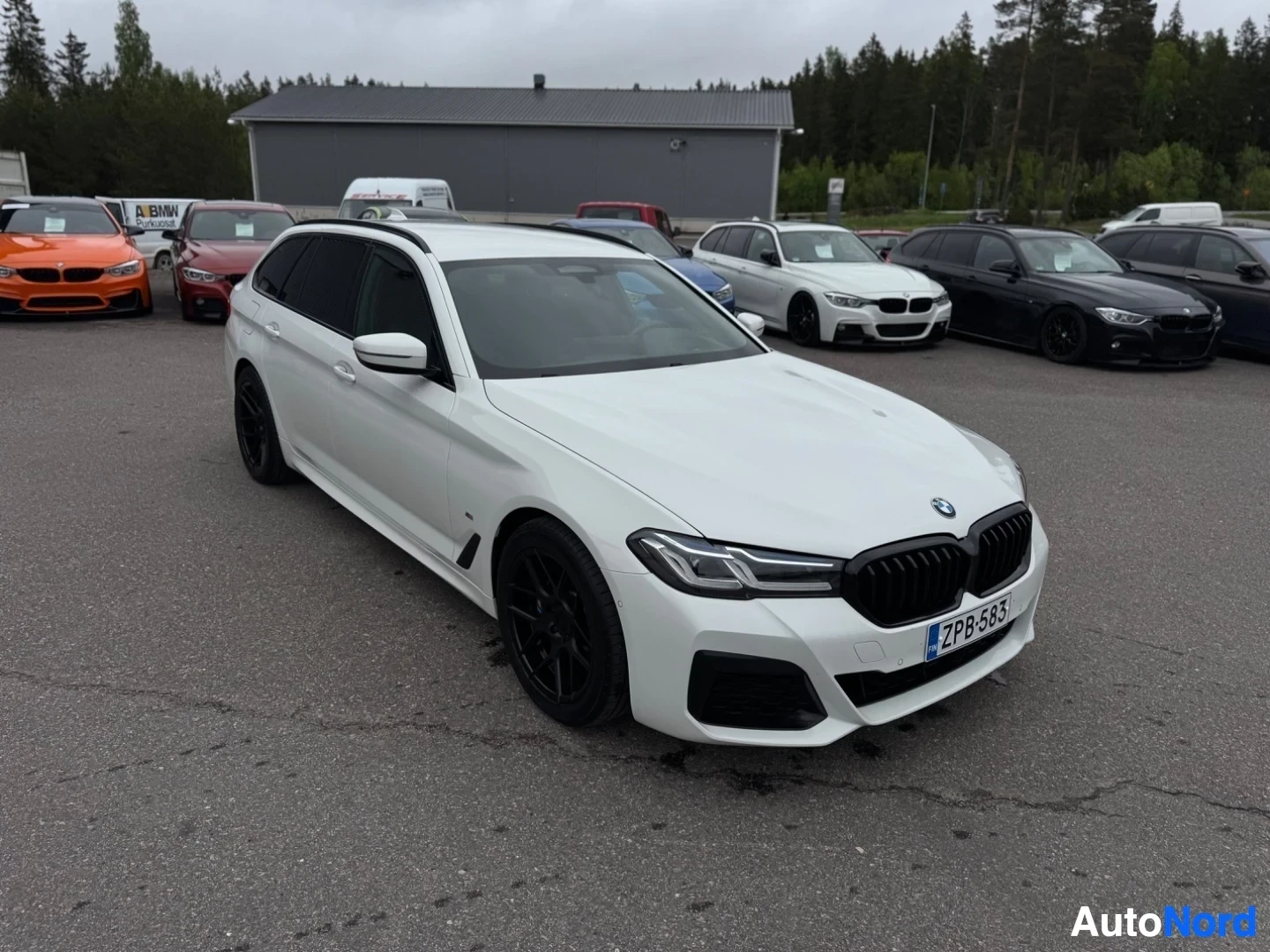 Etuviisto, nokka vasemmalle: Valkoinen farmariauto pysäköitynä asfalttipihalle muiden autojen eteen. Auto on kuvattu hieman yläviistosta edestäpäin, nokka vasemmalle. Rekisterikilp