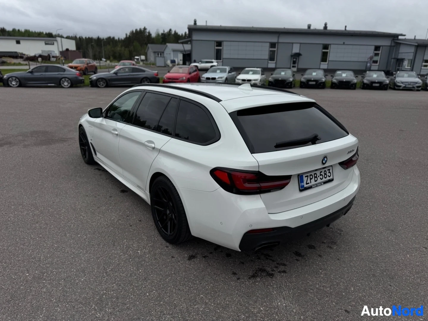 Takaviisto: Valkoinen farmariauto pysäköitynä asfalttipihalle, takapää vasemmalle ja perä kohti kameraa. Auto on kuvattu hieman yläviistosta. Rekisterikilpi on näky